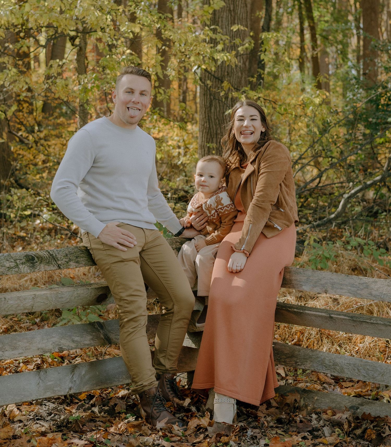 A couple sits together on a wooden fence surrounded by autumn foliage in a romantic outdoor photoshoot.