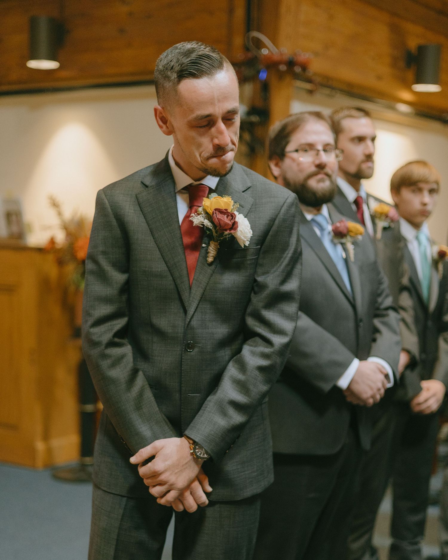 Groomsmen in dark suits stand in a line at a rustic wooden venue with overhead lighting.