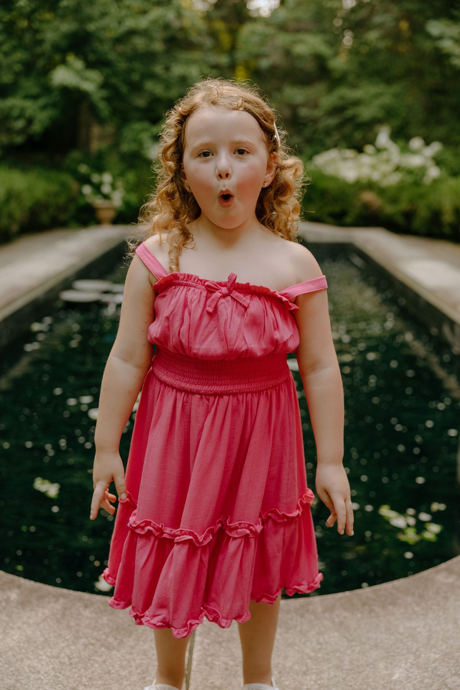 Person in pink ruffled dress poses playfully by circular water feature in garden.