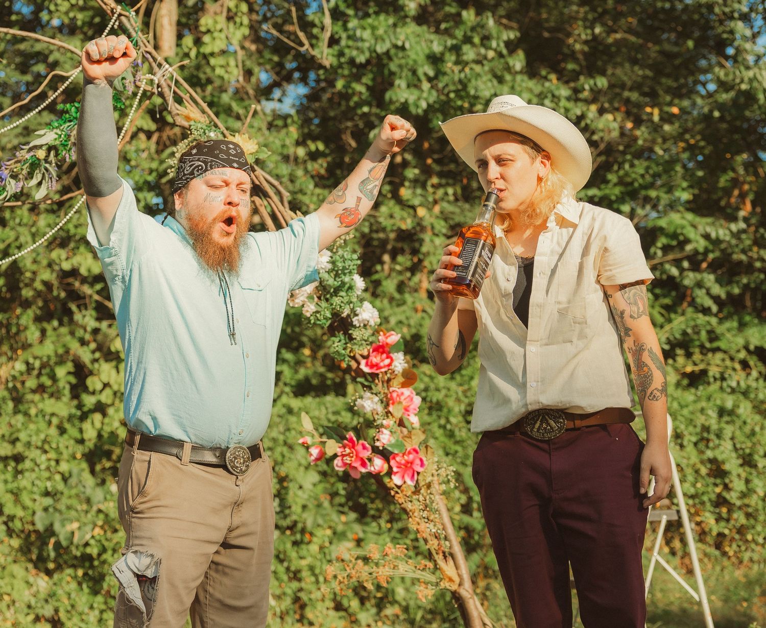 Two older people laugh and smile while looking at pink flowers in a sunny garden setting.
