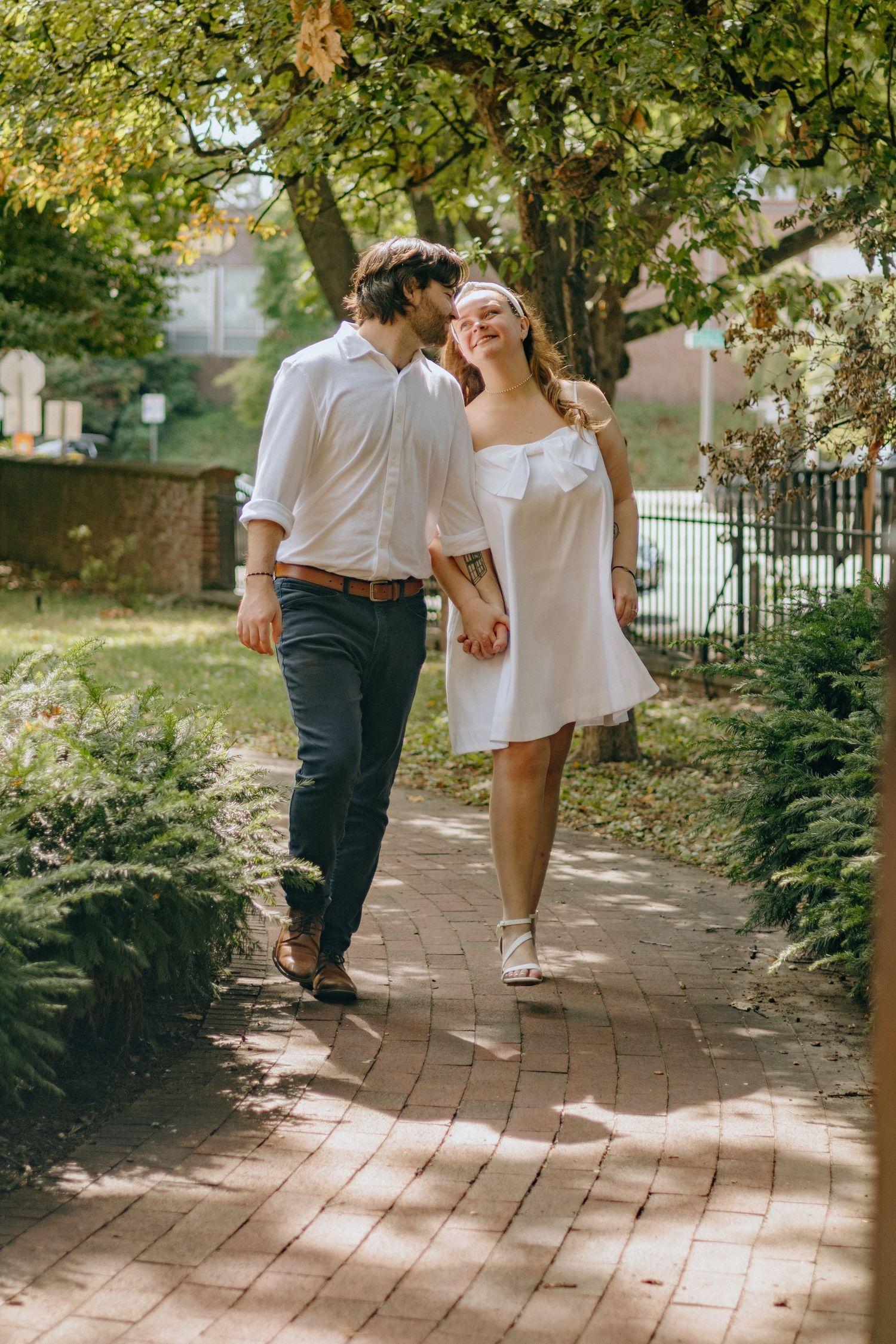 A couple walks hand in hand along a brick pathway under the dappled sunlight filtering through green trees.