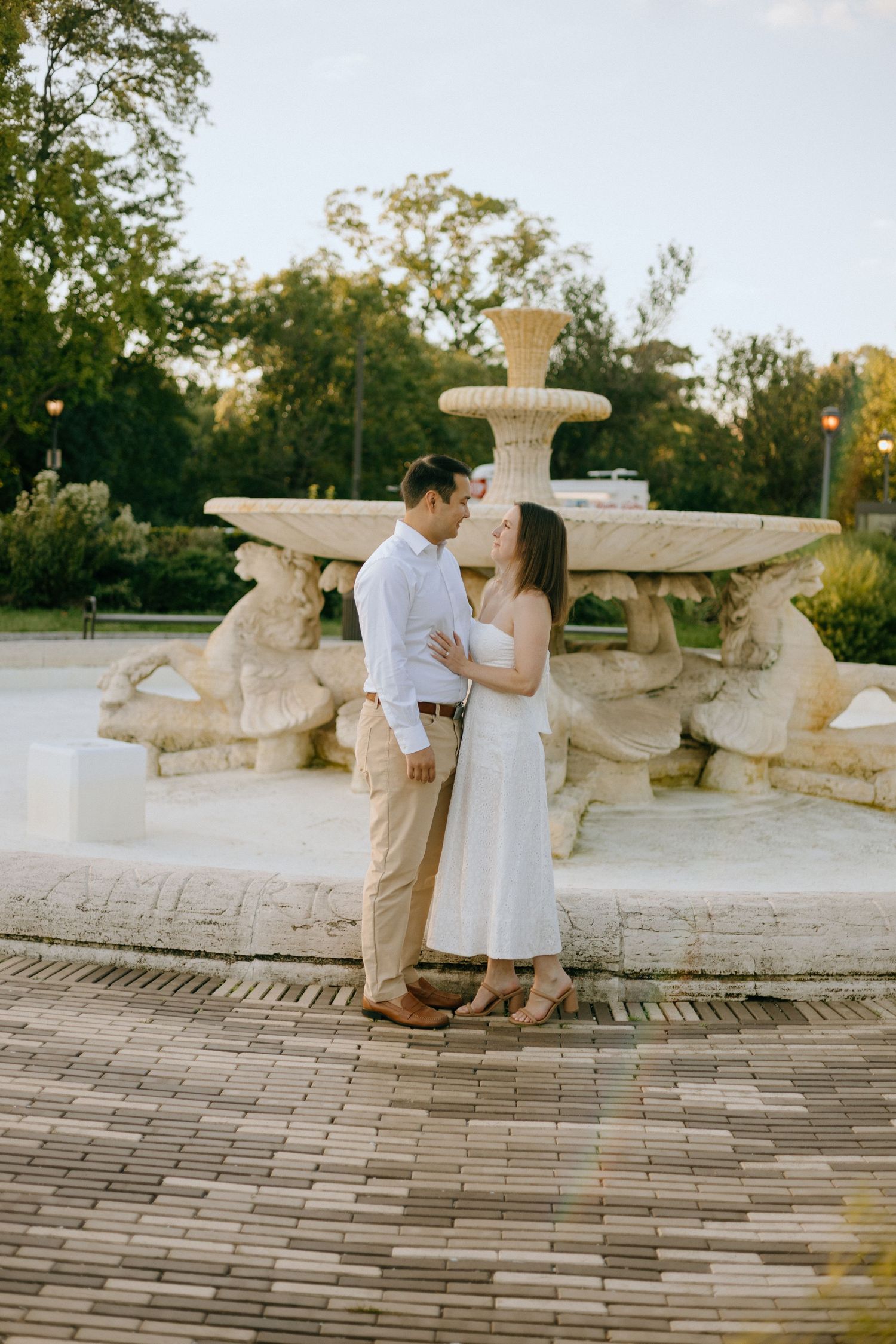 A couple shares a romantic moment near an ornate stone fountain in a garden setting during golden hour.