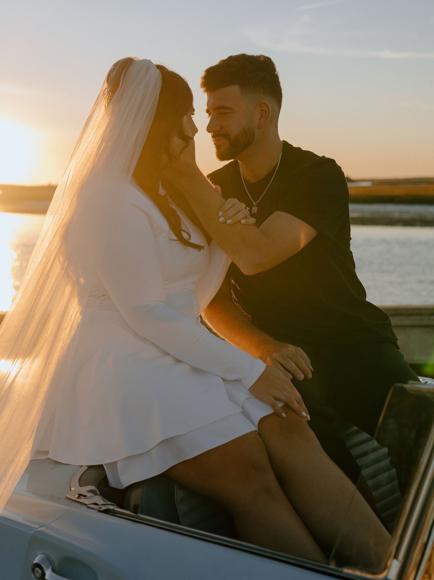 A romantic sunset engagement photo of a couple sitting in a vintage car by the water.