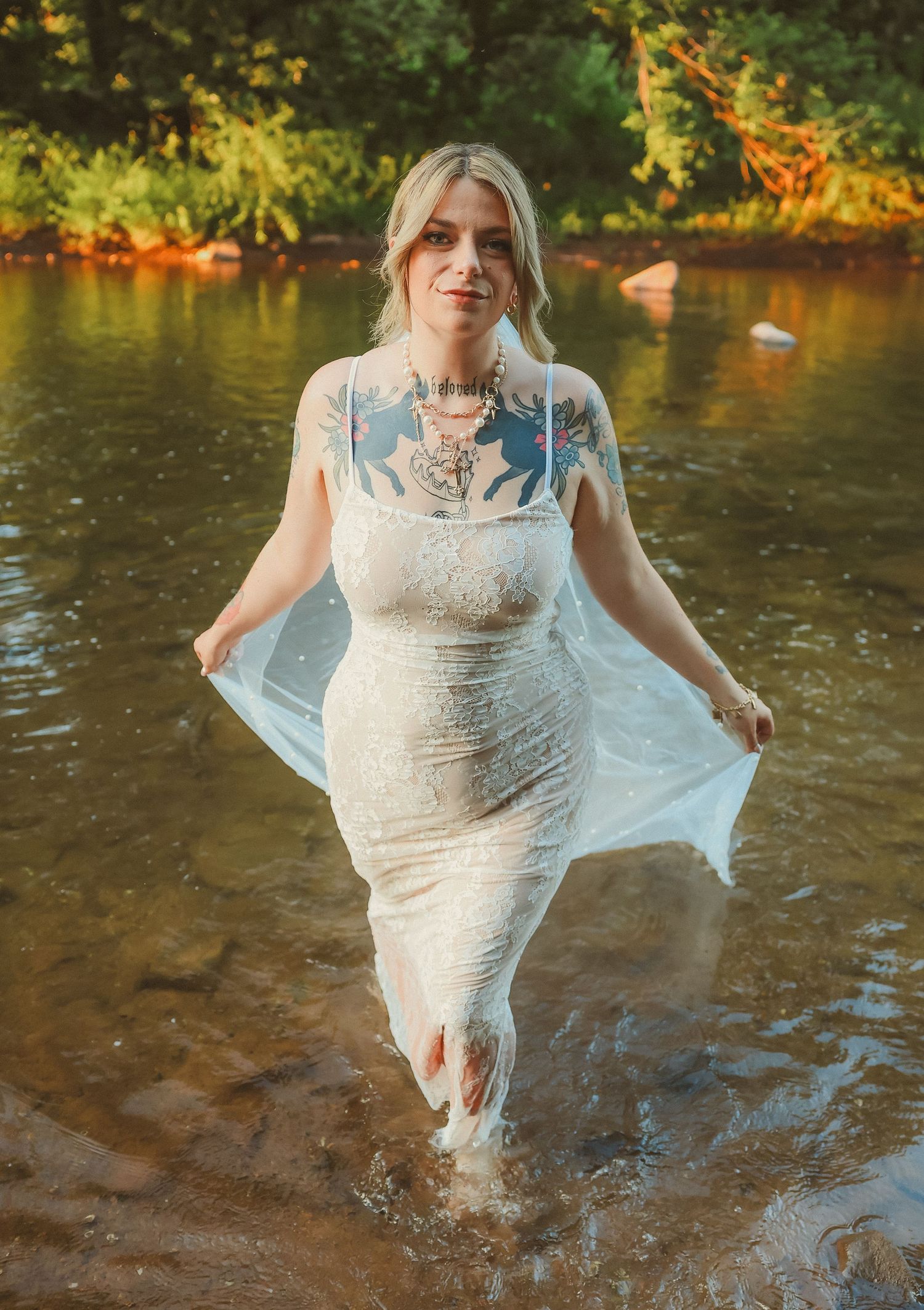 A bride in a lace wedding dress stands in flowing river water with her veil floating behind her.