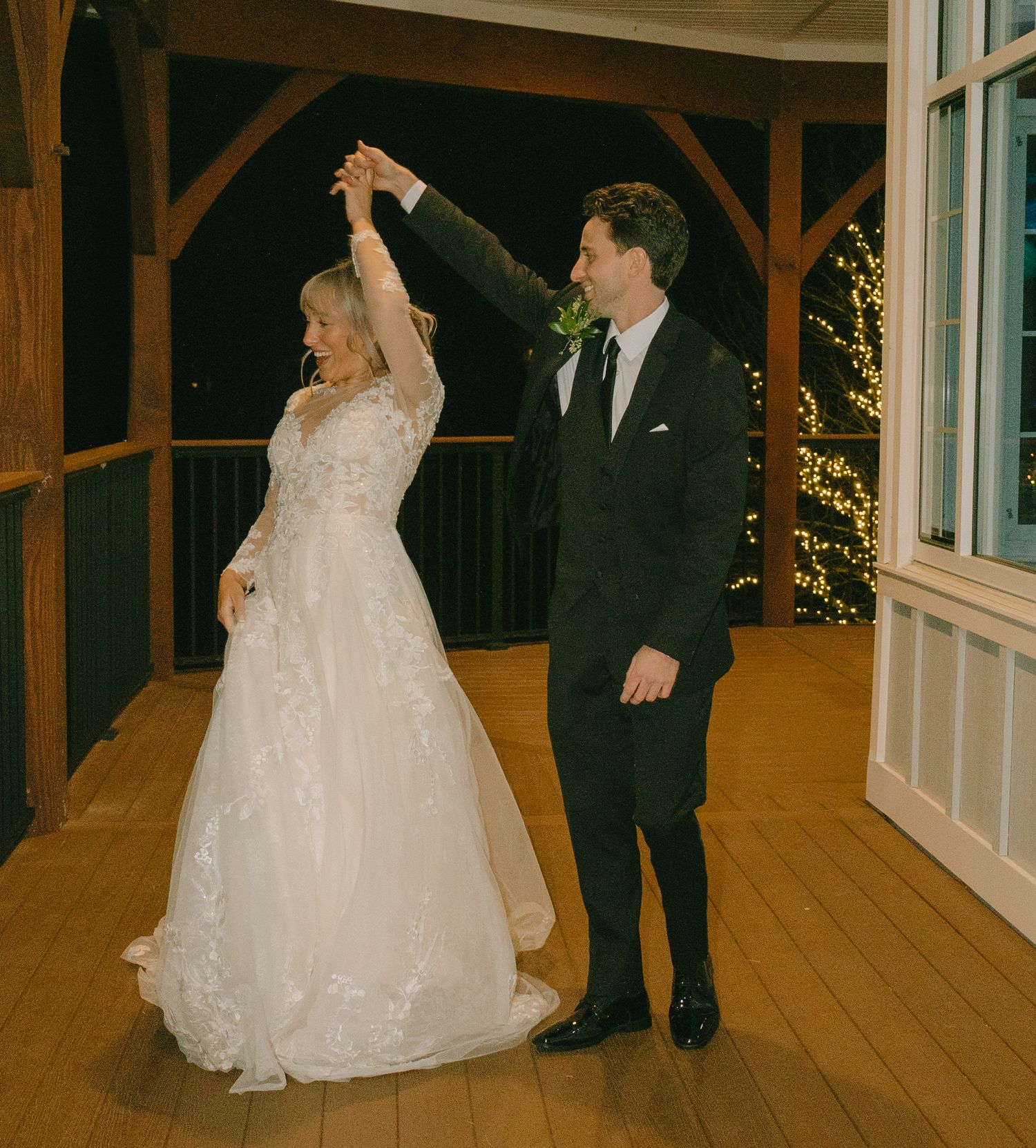 Wedding couple dances together on wooden floor in rustic venue with twinkling lights.