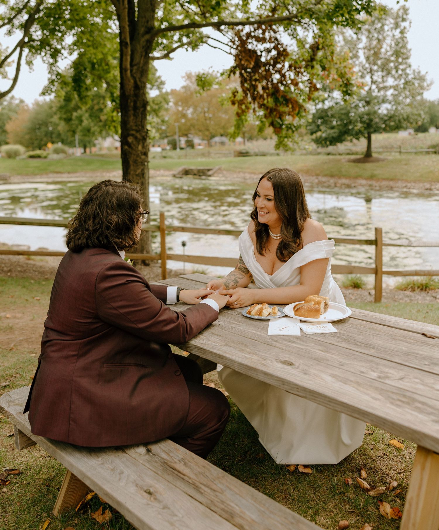 Two people sit at opposite ends of a wooden picnic table in a park setting on a sunny autumn day.