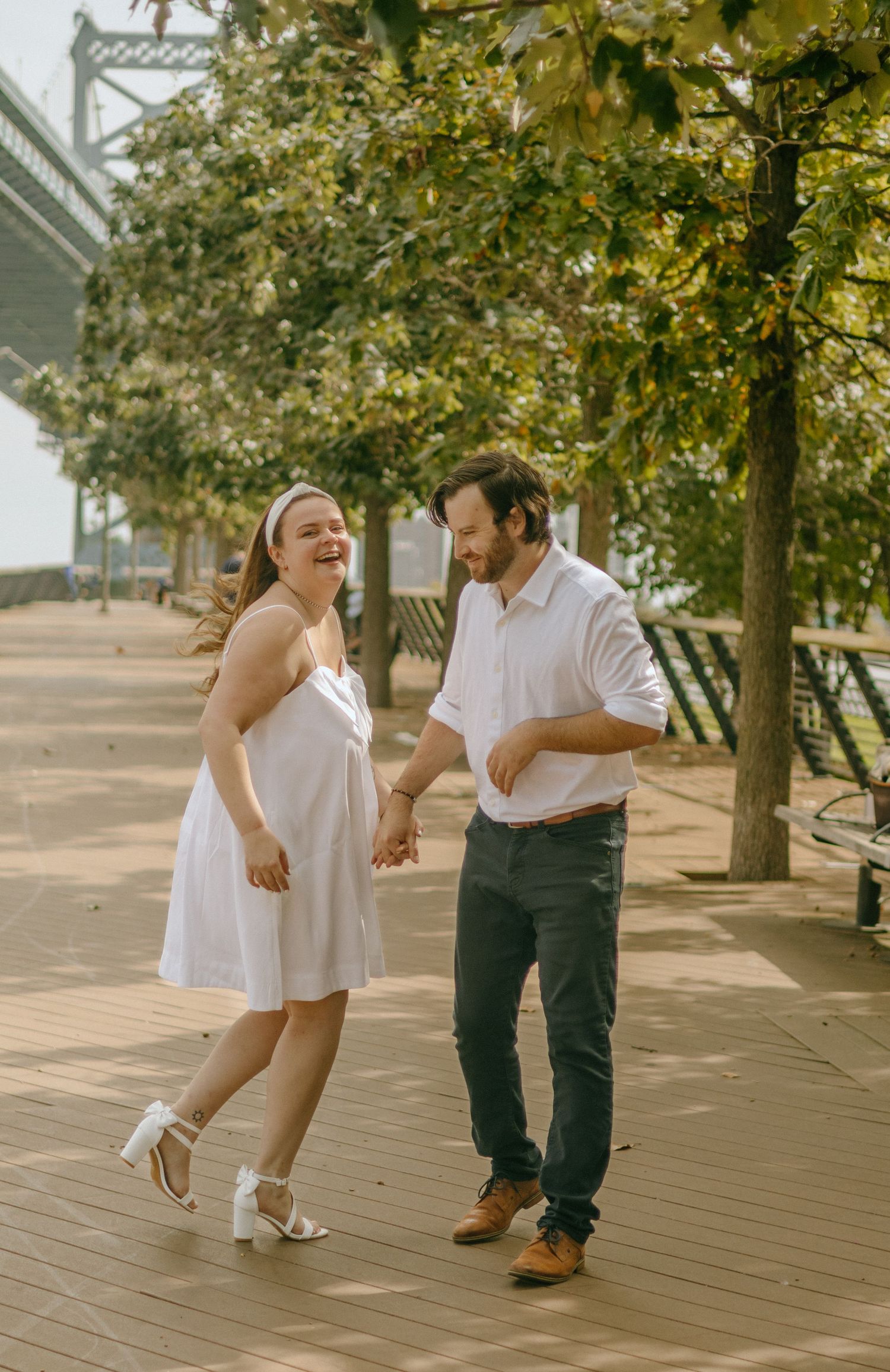 A couple in white and dark clothing dance playfully together on a wooden deck under autumn trees.