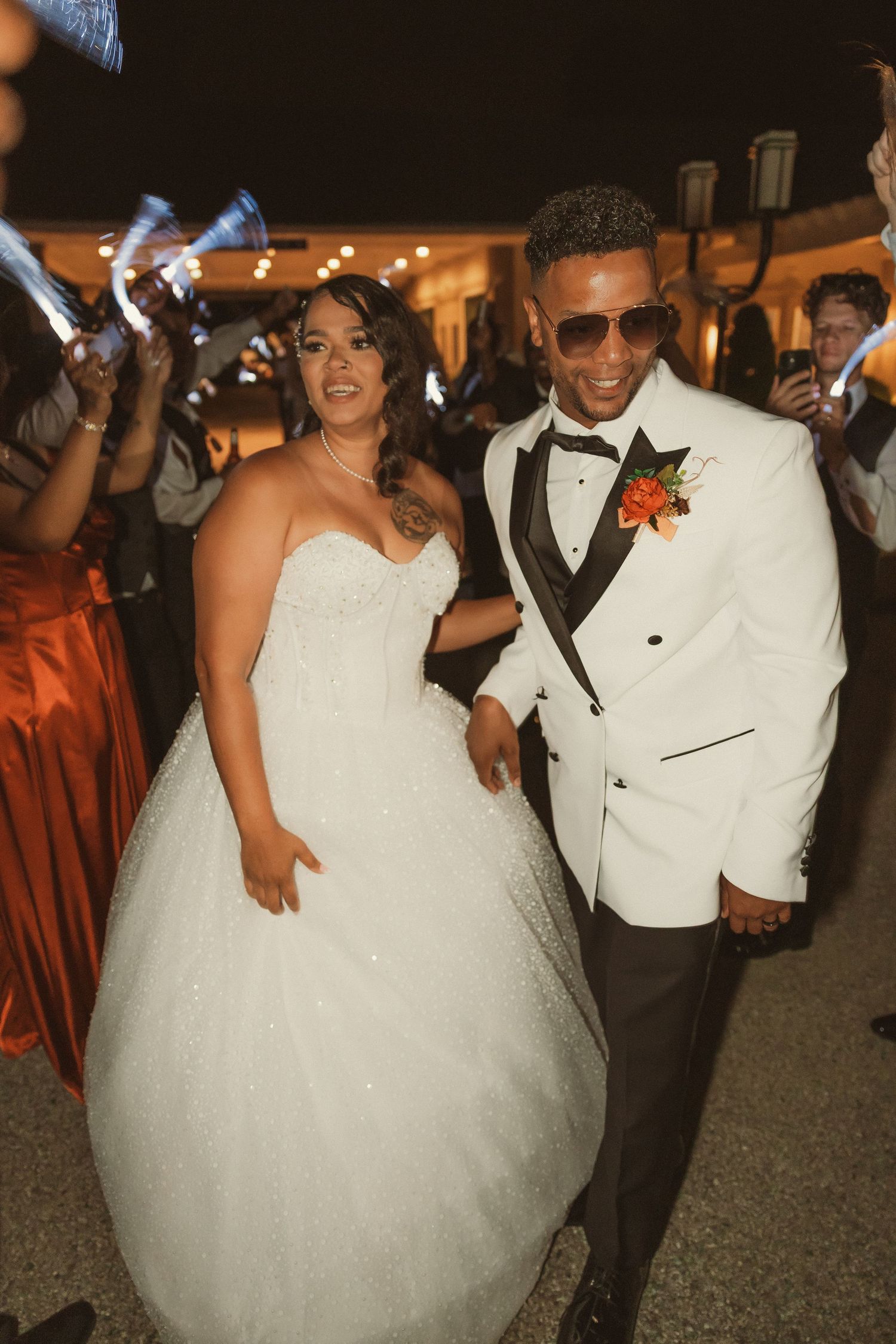 Newlyweds in white attire dance together at an evening reception with guests.