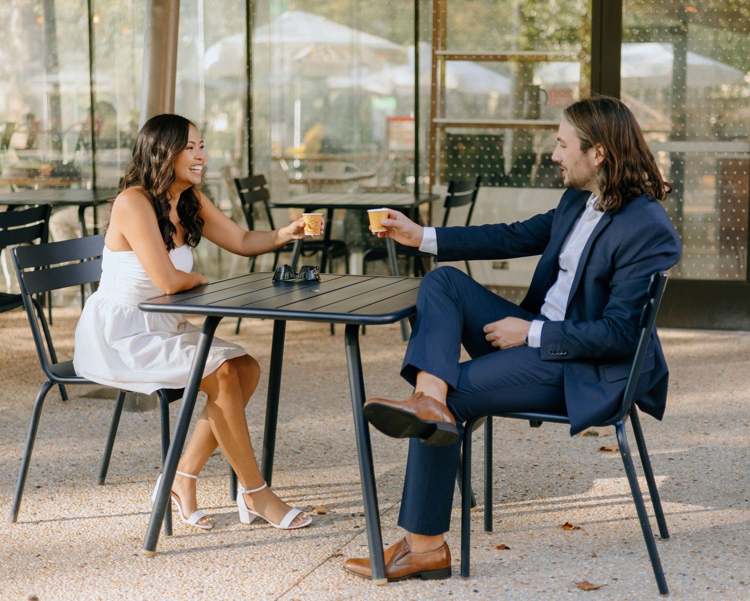 People in formal attire sit at outdoor tables having an animated conversation in a garden setting.