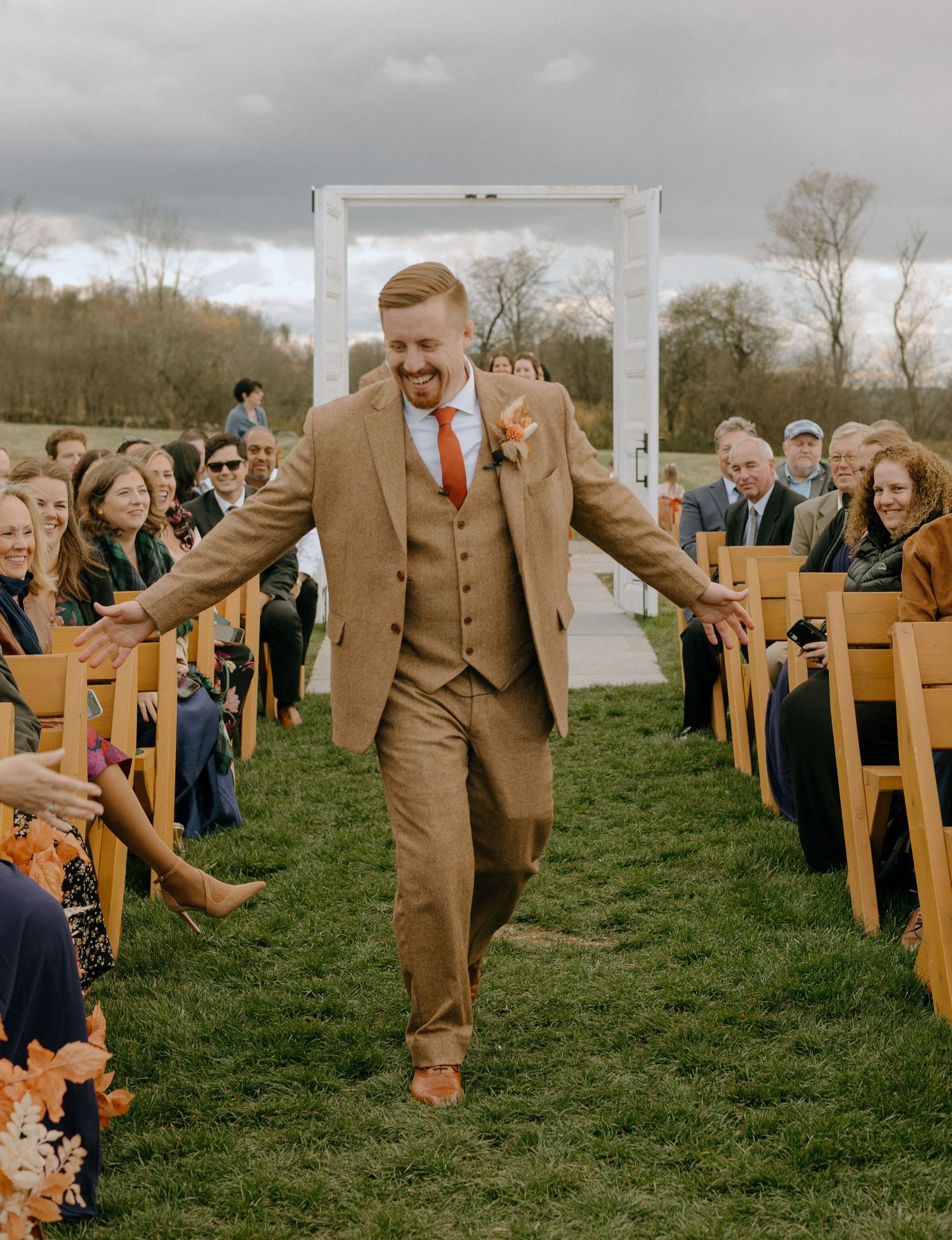 Groomsmen in brown suits playfully walking down an outdoor aisle lined with wooden chairs.