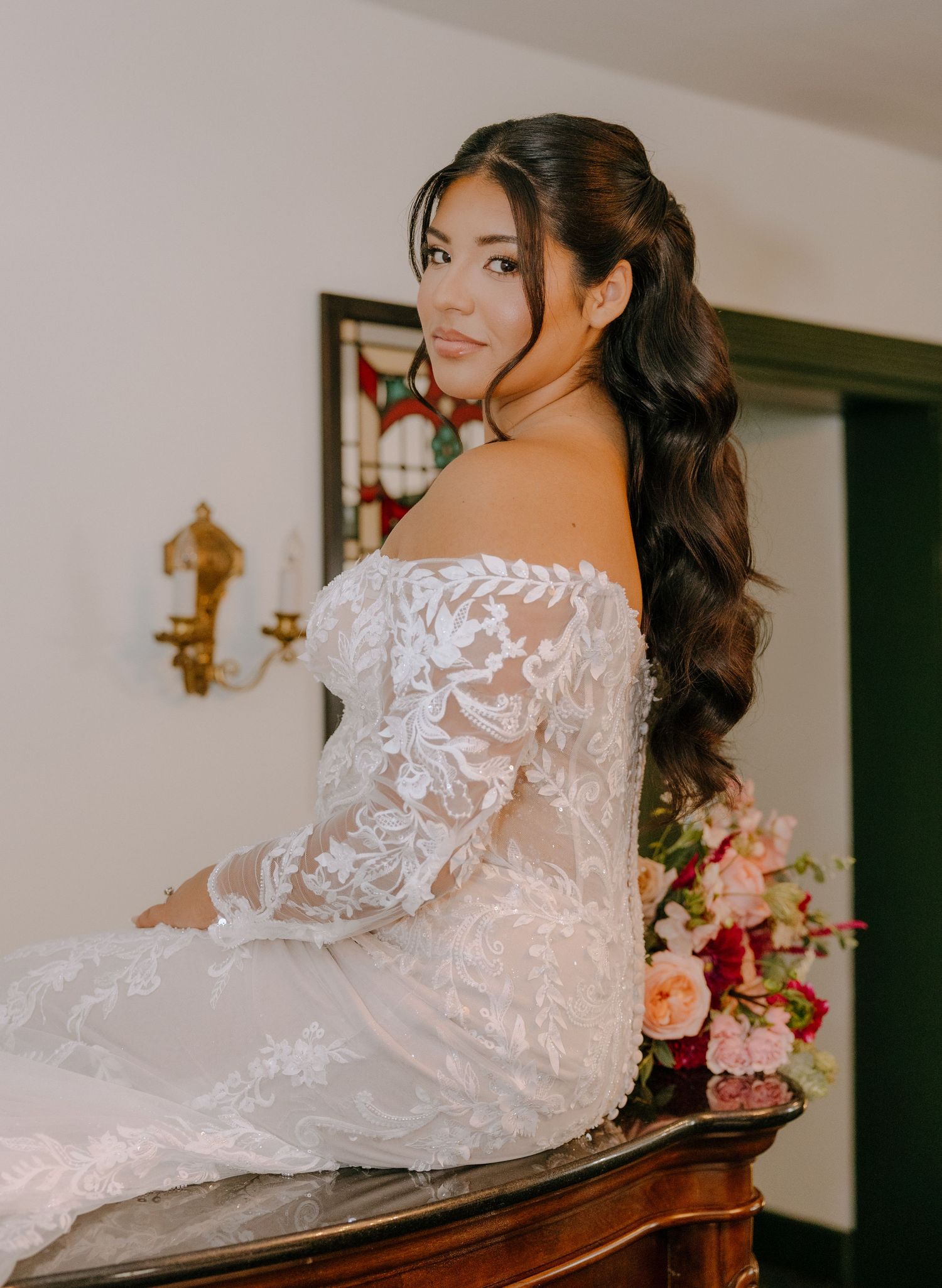 Bride in detailed lace gown poses gracefully on antique furniture.