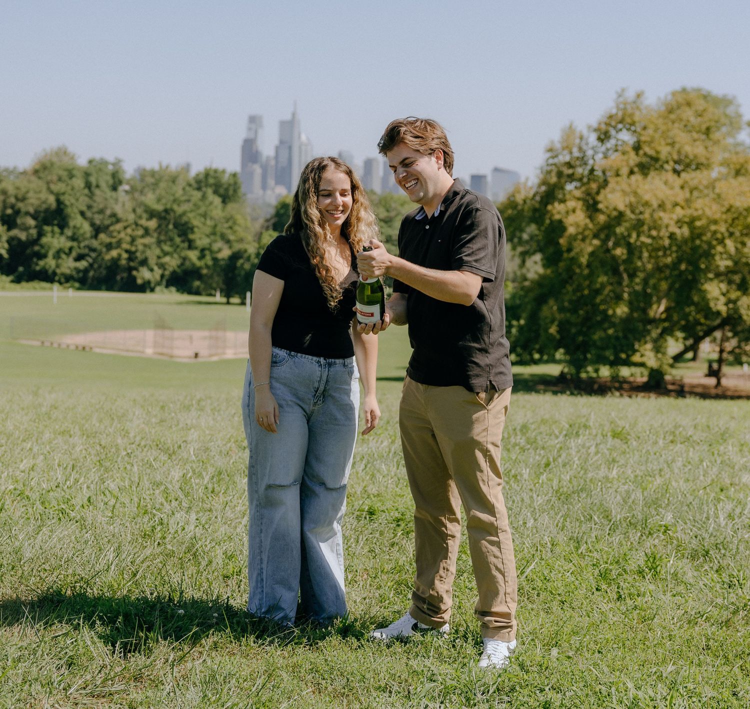 A couple enjoys a romantic picnic in a grassy park with the city skyline visible in the background.