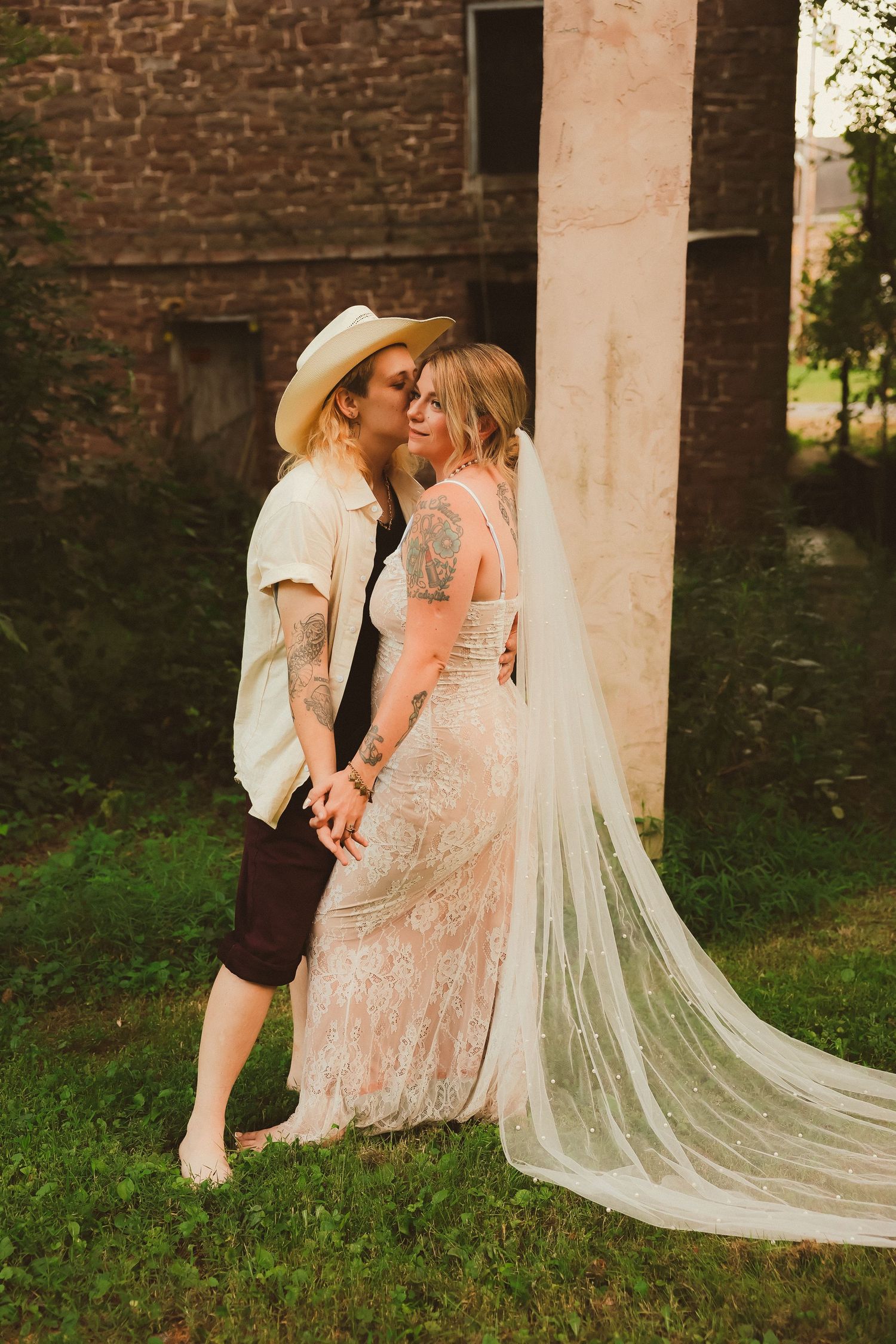 A romantic couple poses against a brick wall, with one wearing a white hat and the other in a lace wedding dress with veil.