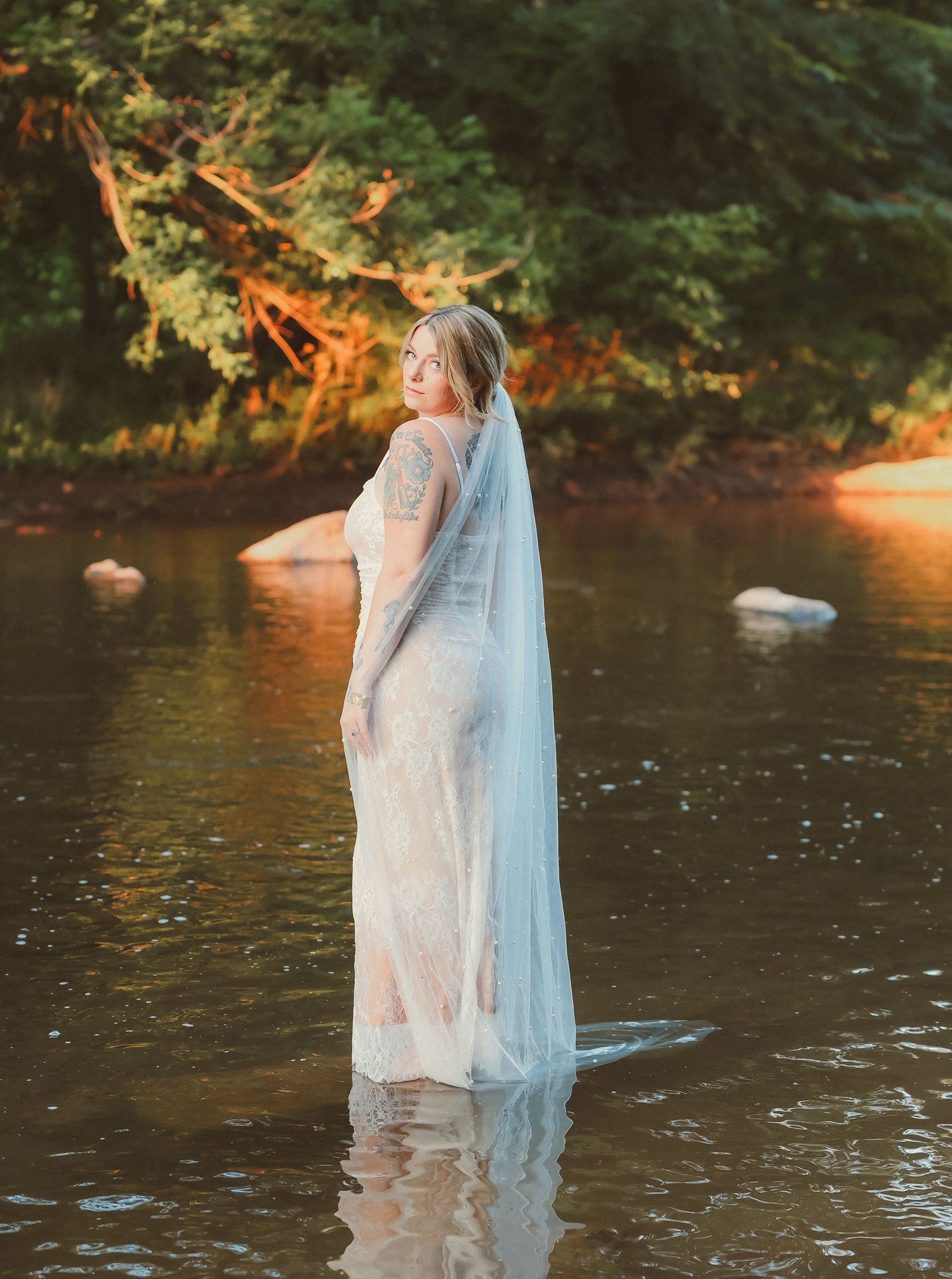 A dreamy bridal portrait showing a person in a lace dress standing in a river during sunset.