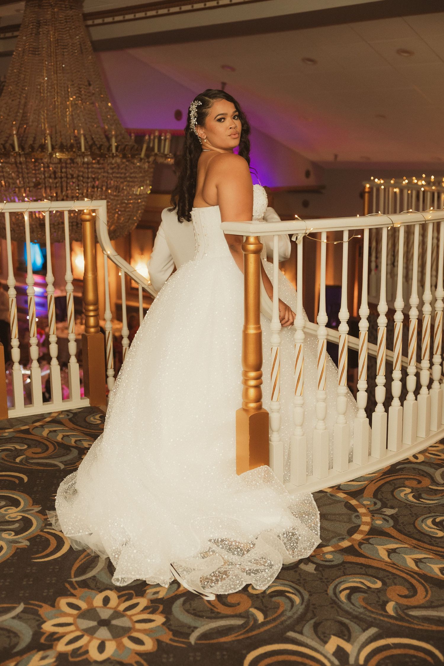 A bride in a flowing white gown poses near ornate wooden railings in an elegant venue.