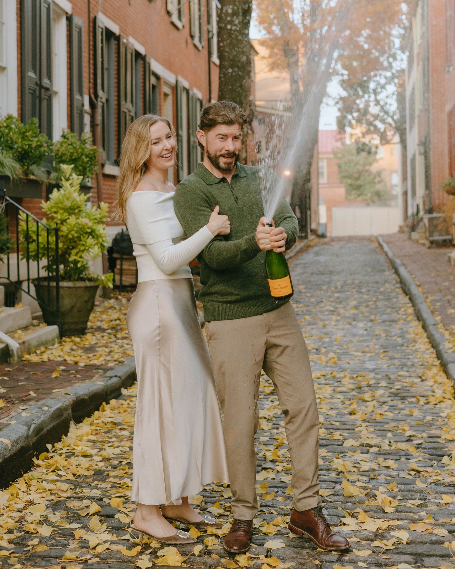 A couple sprays champagne while celebrating on a scenic autumn street lined with brick buildings.