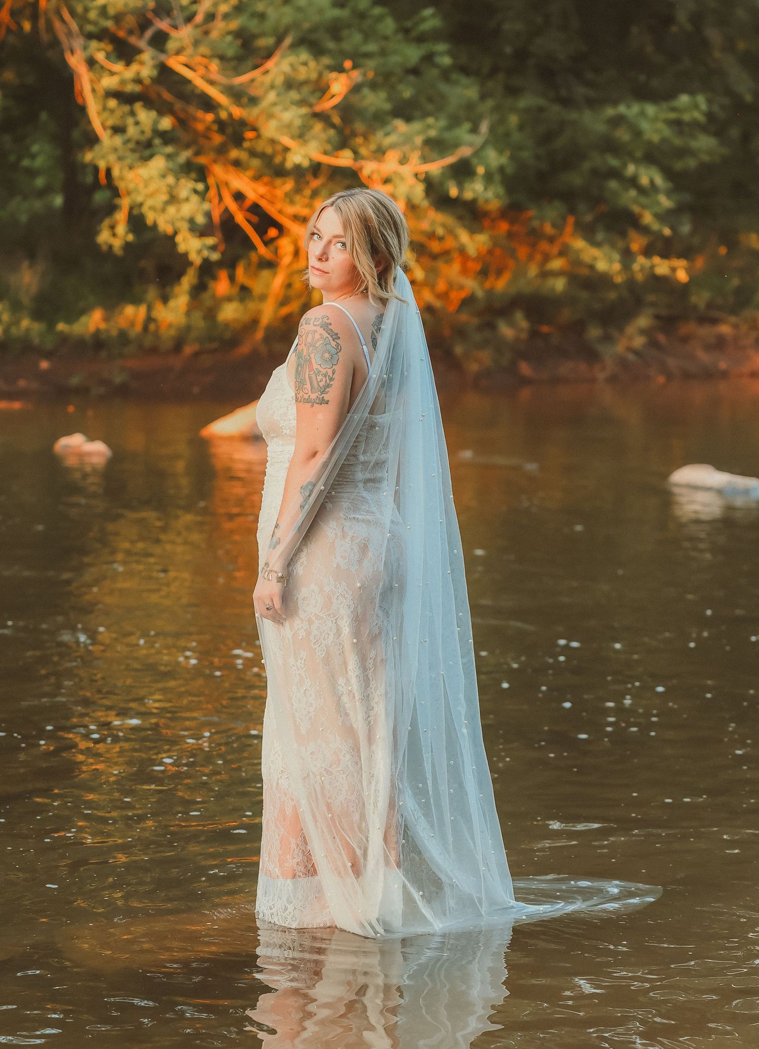 A person wearing a lace wedding dress wades through river water at sunset.