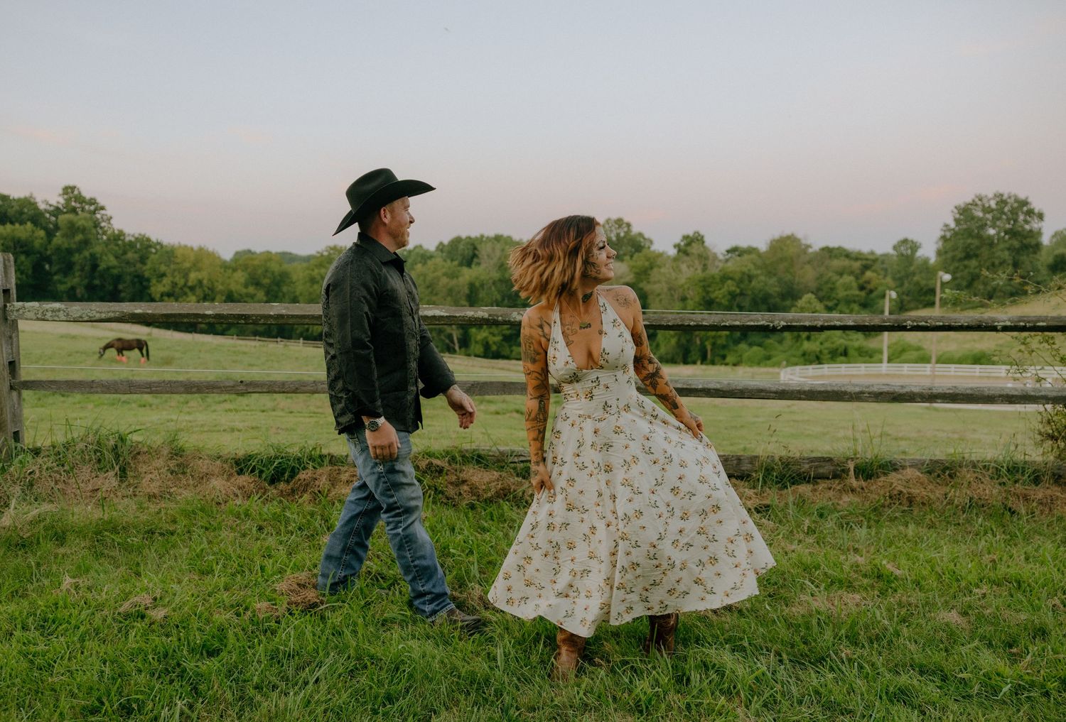 A couple dances together in a green field with wooden fencing and trees in the background.