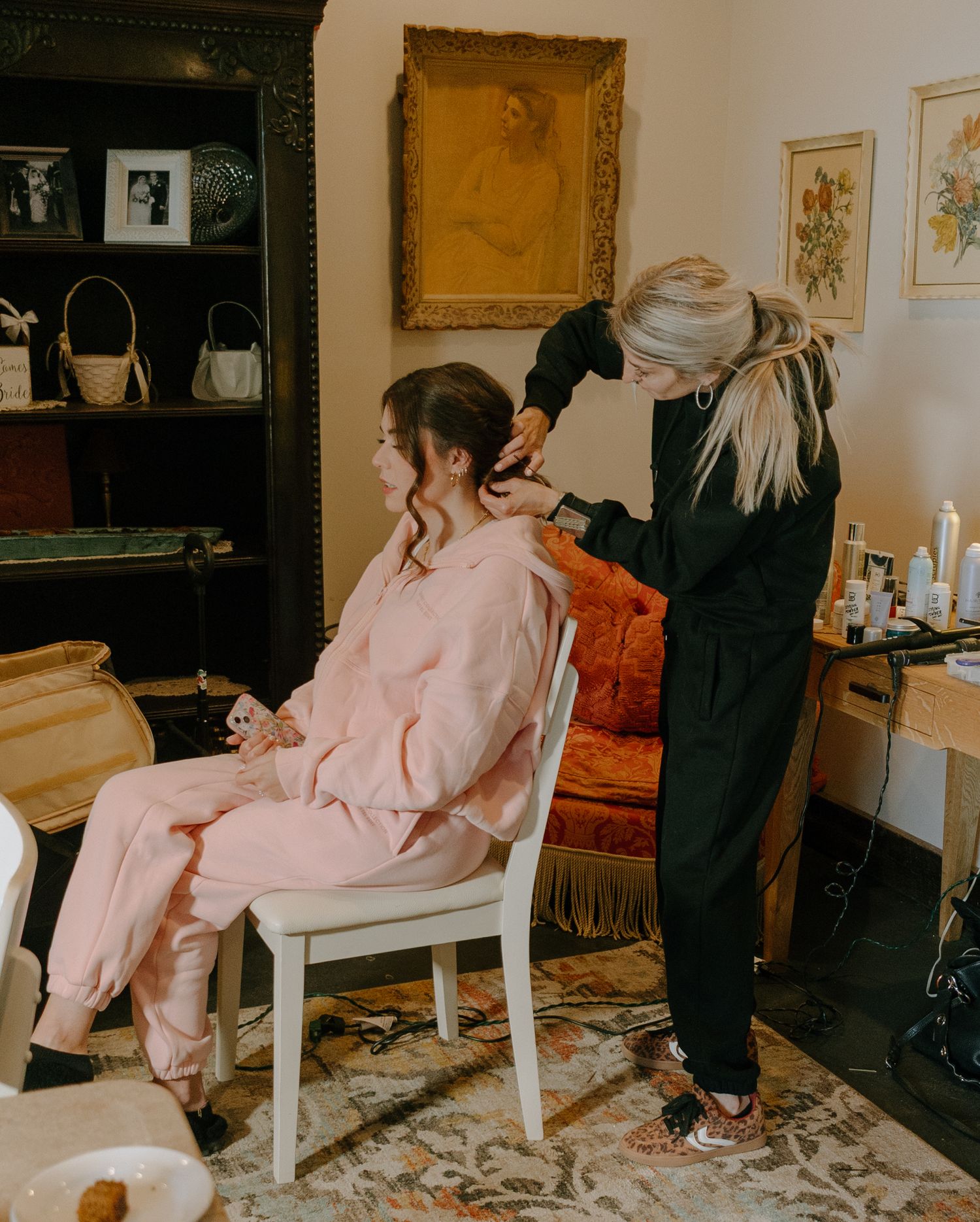 A person in a pink robe sits on a white chair while getting ready in a room with framed artwork on the walls.