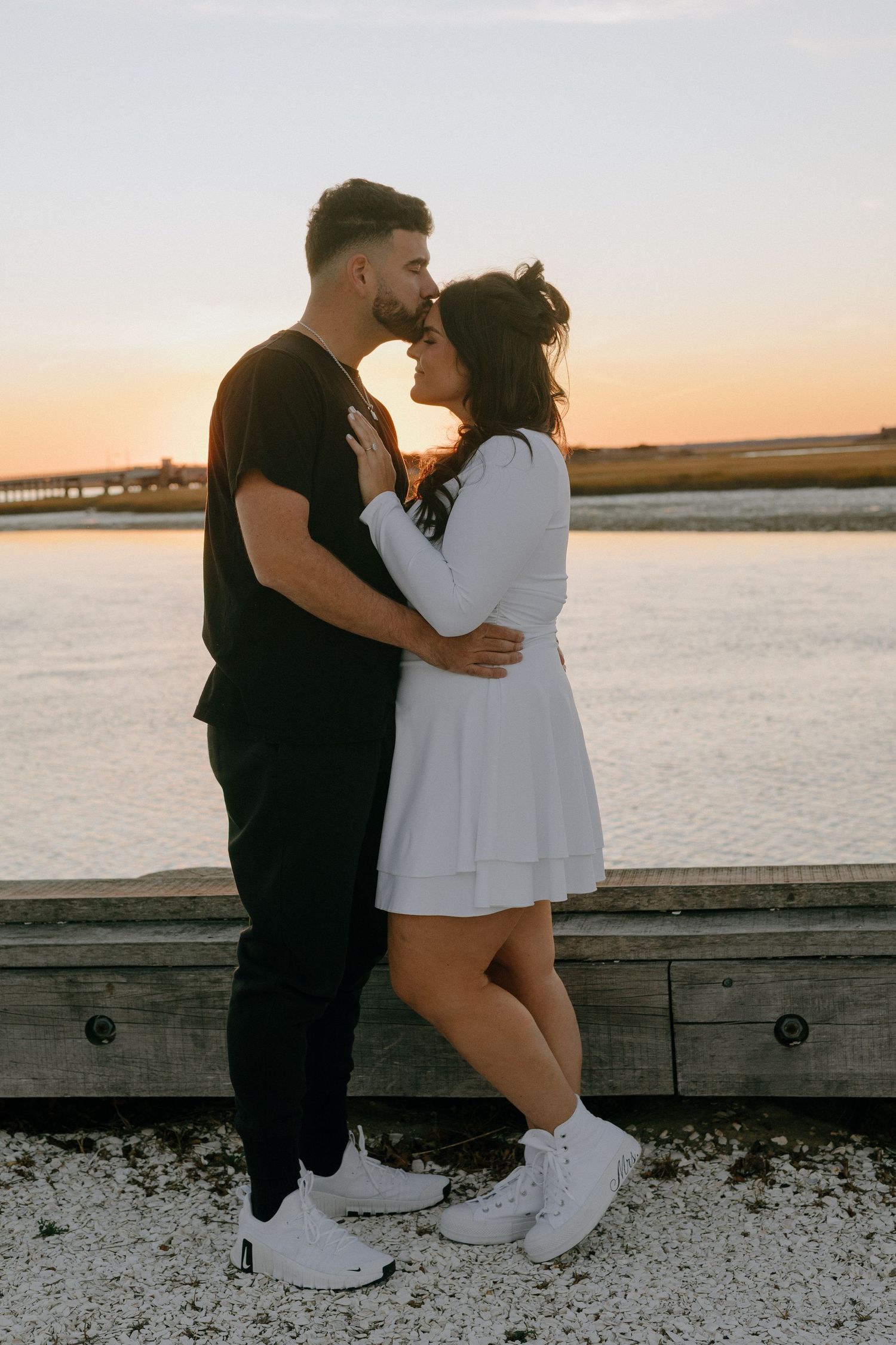 Couple in black and white outfits sharing romantic moments by the waterfront at sunset.