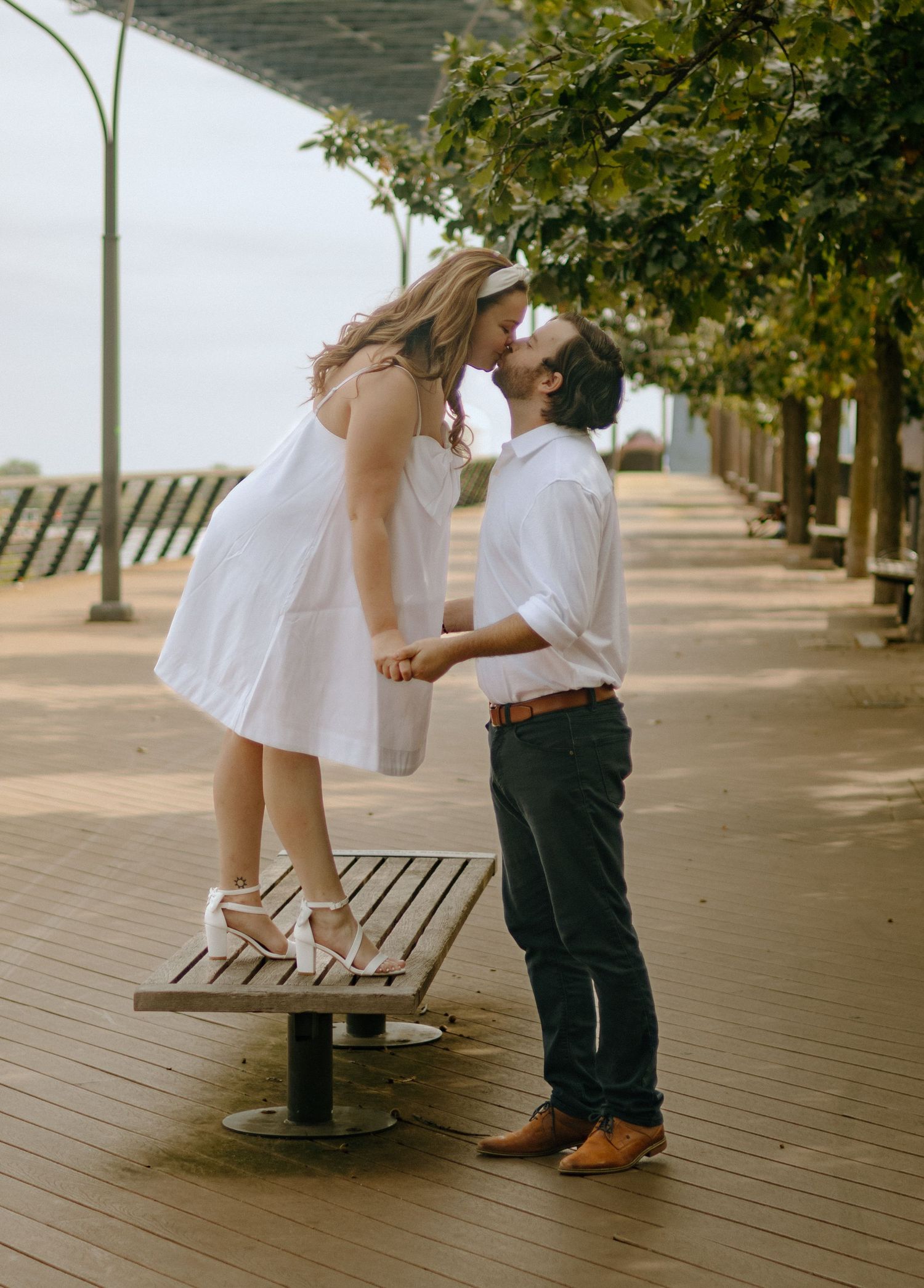 A romantic couple embraces on a wooden boardwalk lined with trees on a sunny afternoon.
