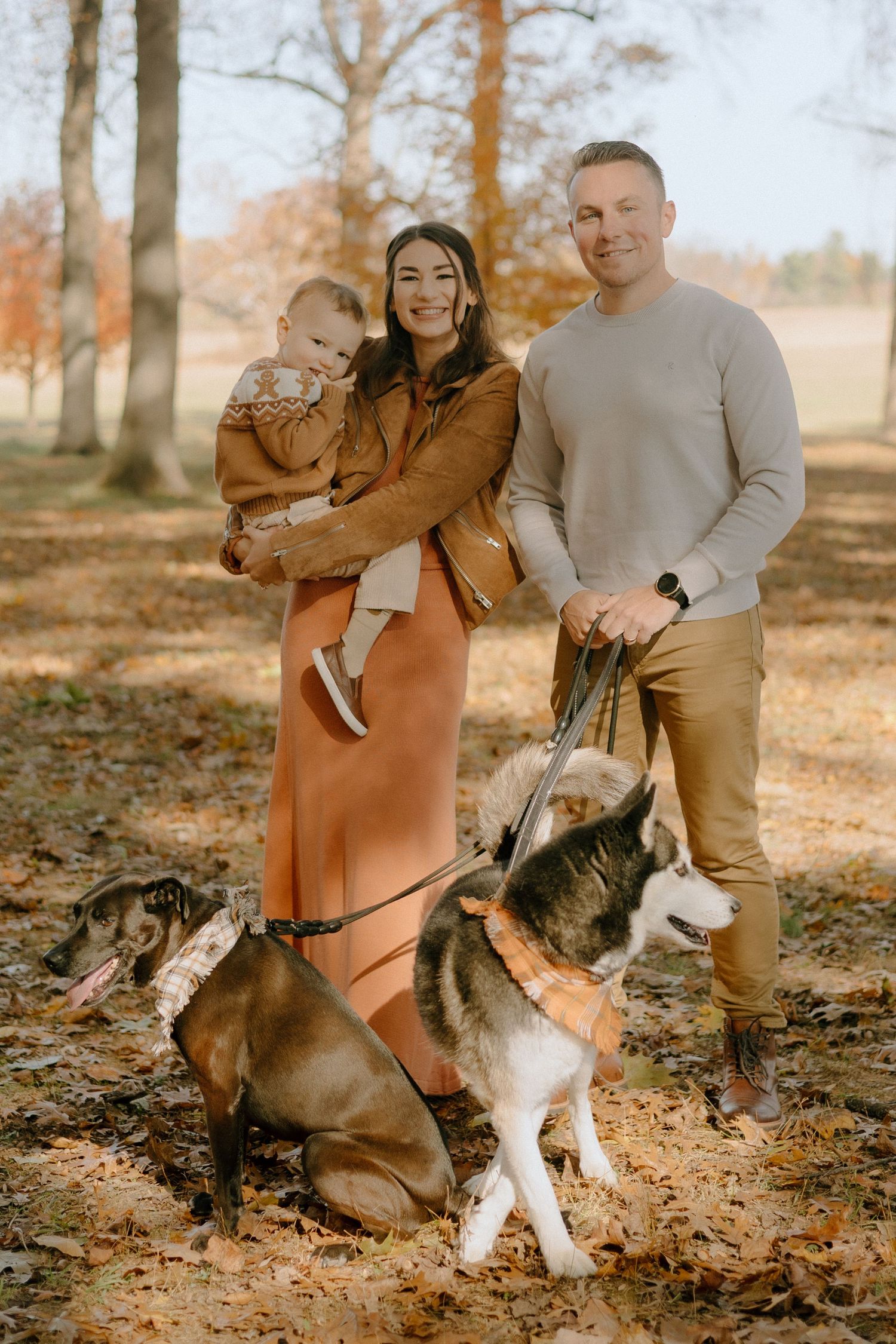 A couple poses with their two dogs in a park setting with autumn leaves scattered on the ground during a photoshoot.