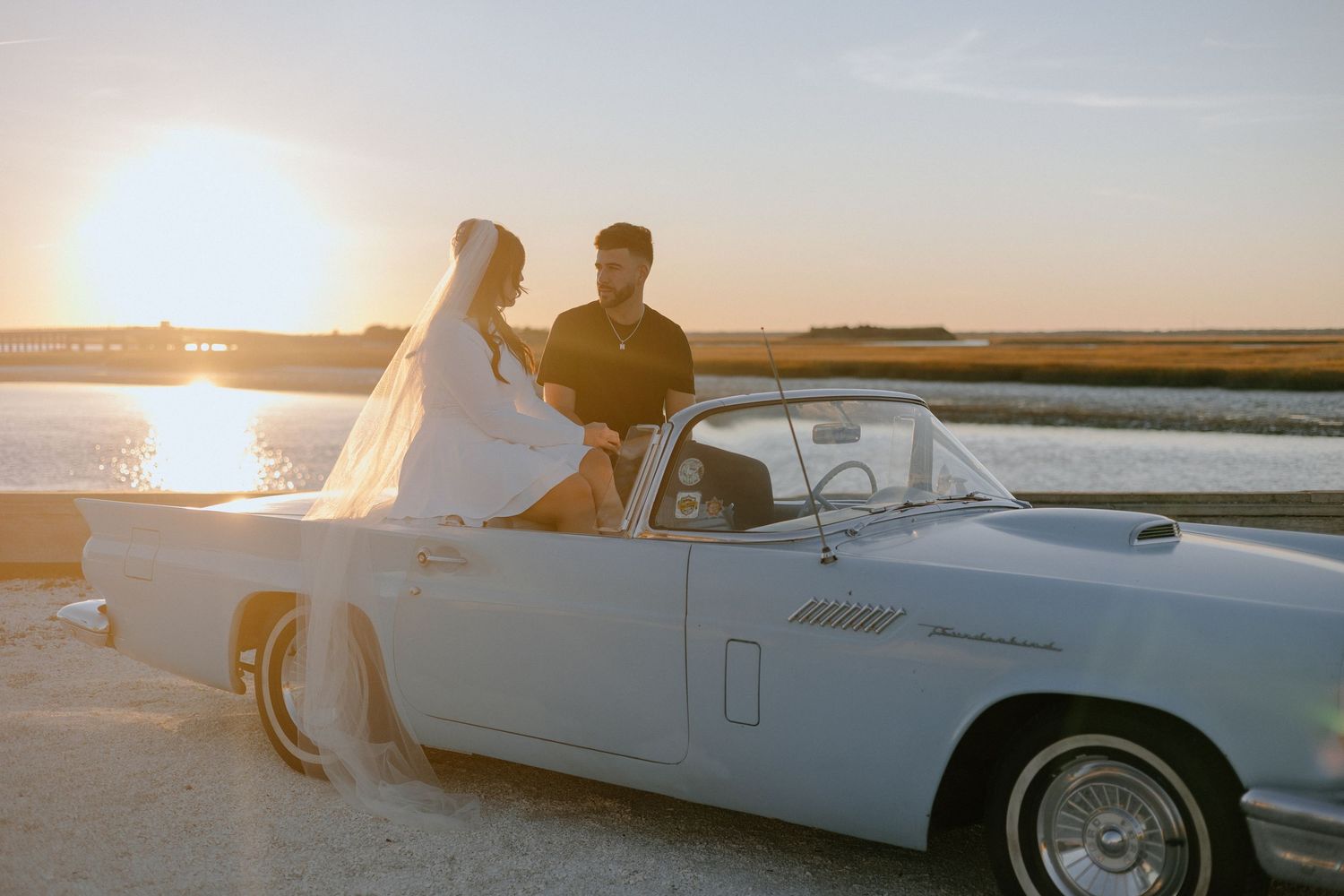 Vintage white convertible parked by waterfront at sunset with couple sitting on car door.