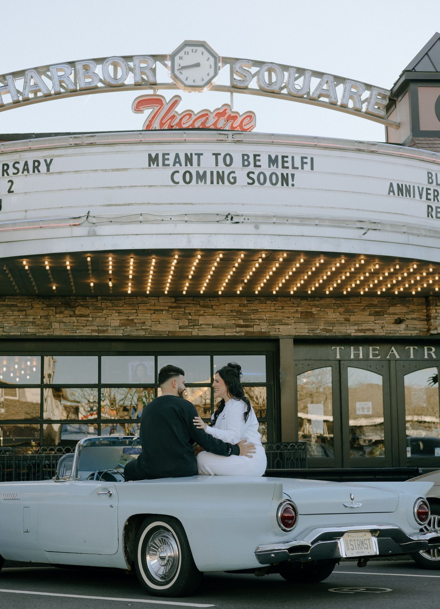 Classic white Thunderbird car parked in front of vintage theater marquee with illuminated lights at dusk.
