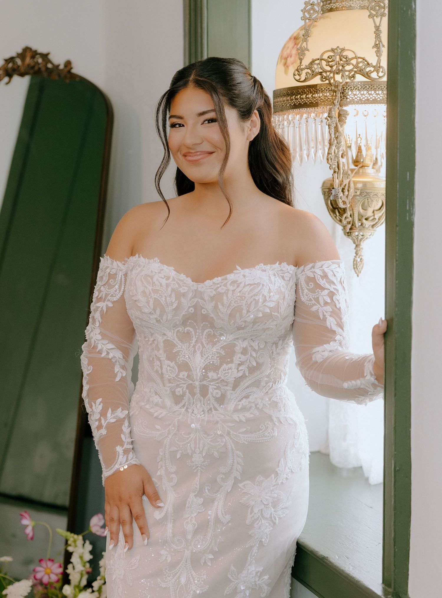 Bride in detailed lace off-shoulder wedding gown poses elegantly indoors.