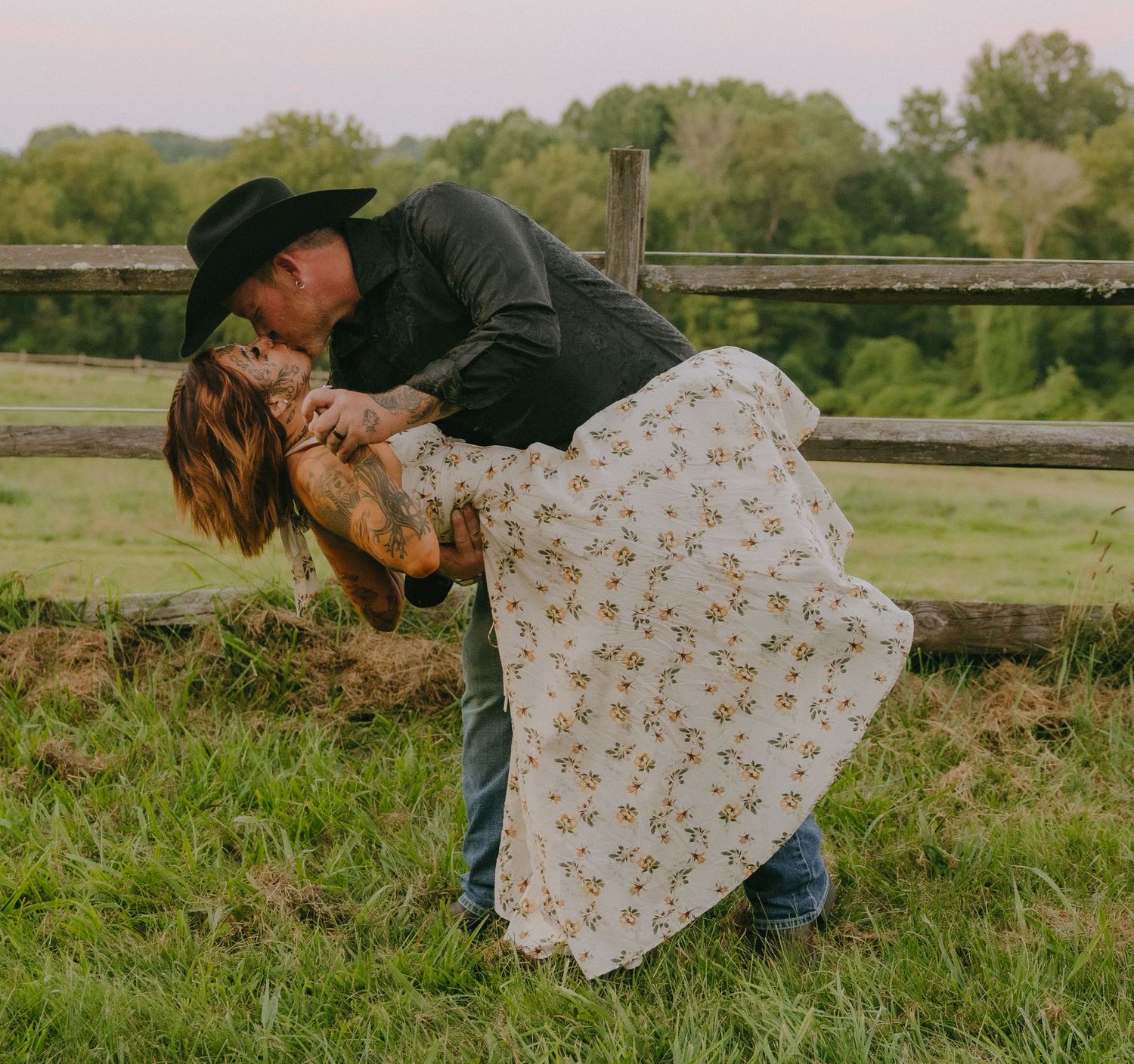 Playful couple poses by wooden fence in rural setting during golden hour.