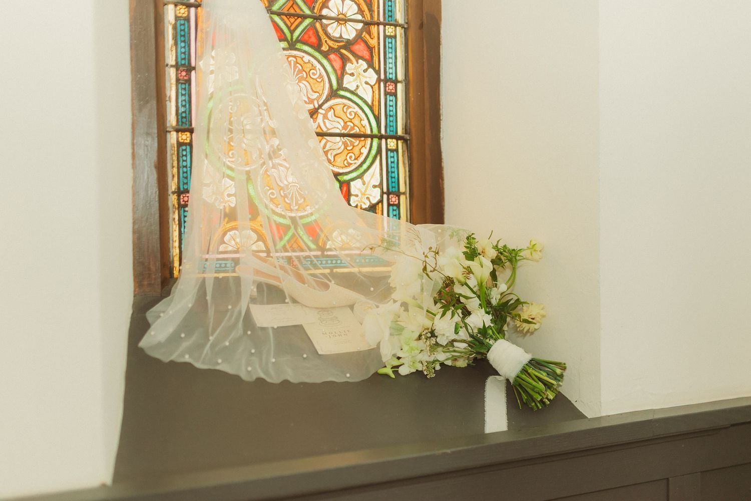 Delicate white bridal veil and bouquet arranged near stained glass window.