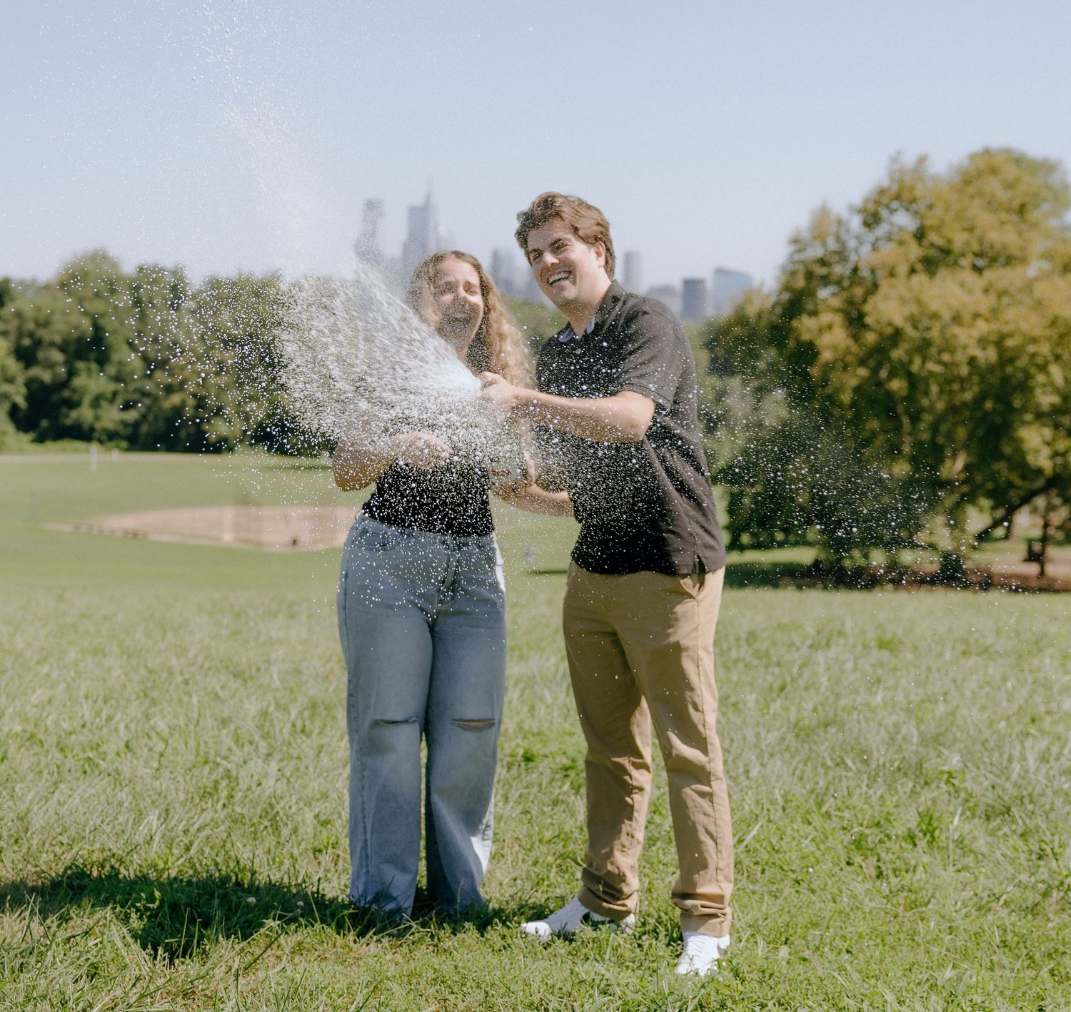 Two people pop a champagne bottle in celebration on a sunny day in an open field.