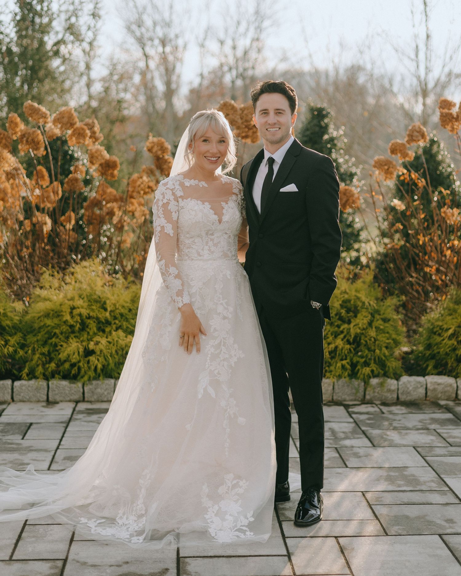 A bride and groom pose together on a stone patio with autumn foliage in the background.