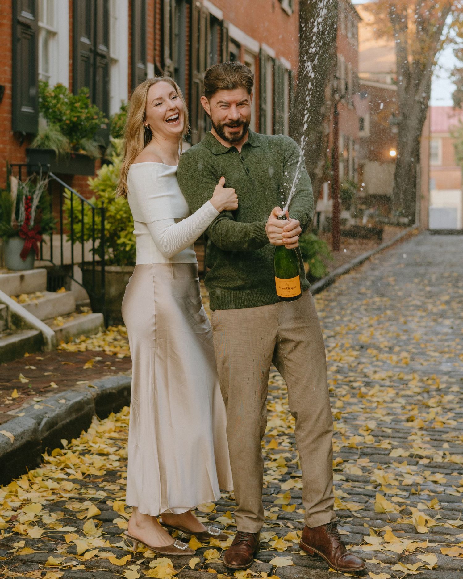 A joyful couple celebrating with champagne on an autumn street lined with brick buildings and fallen leaves.