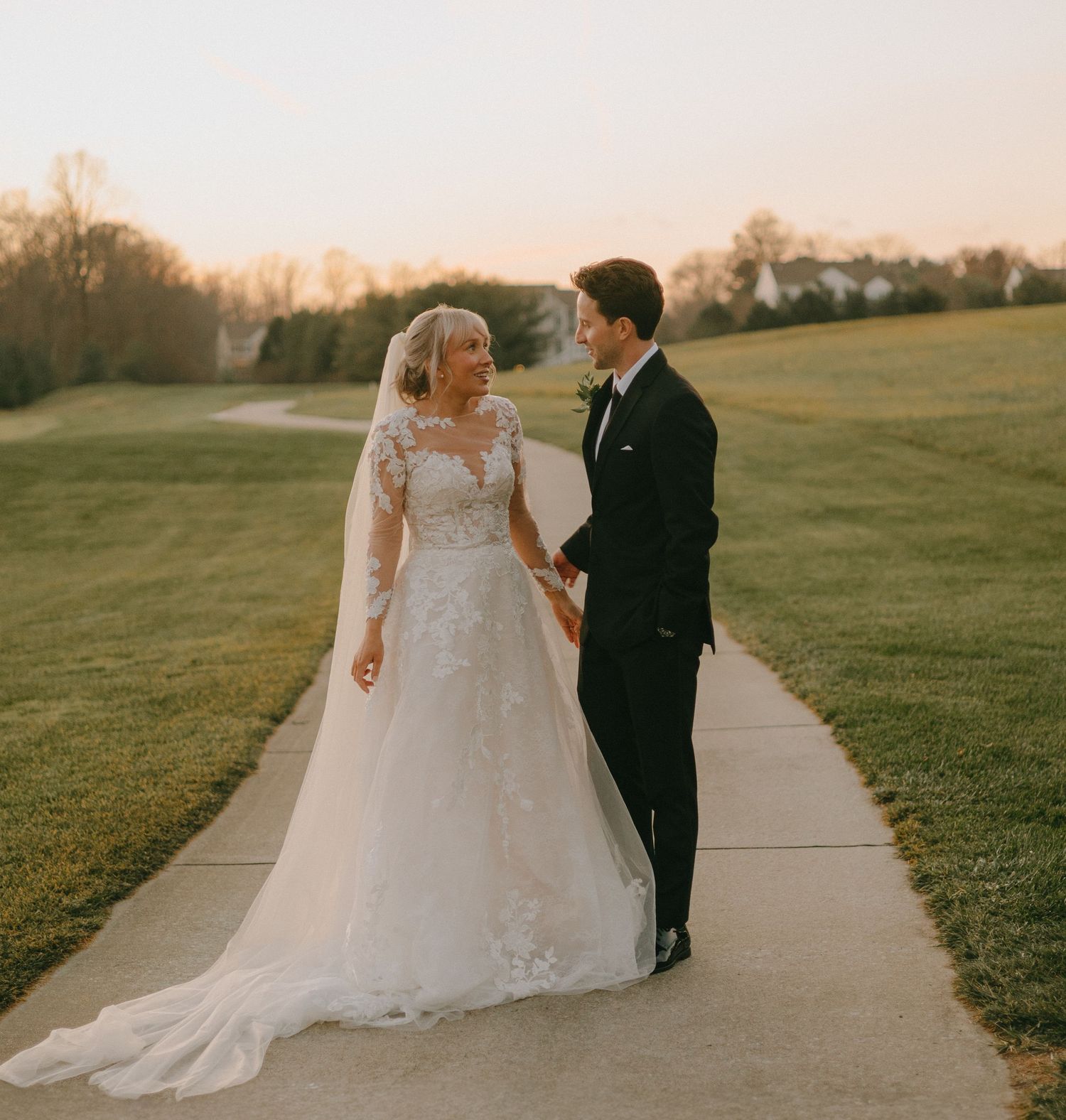 A bride and groom walk together on a pathway during golden hour surrounded by green fields.