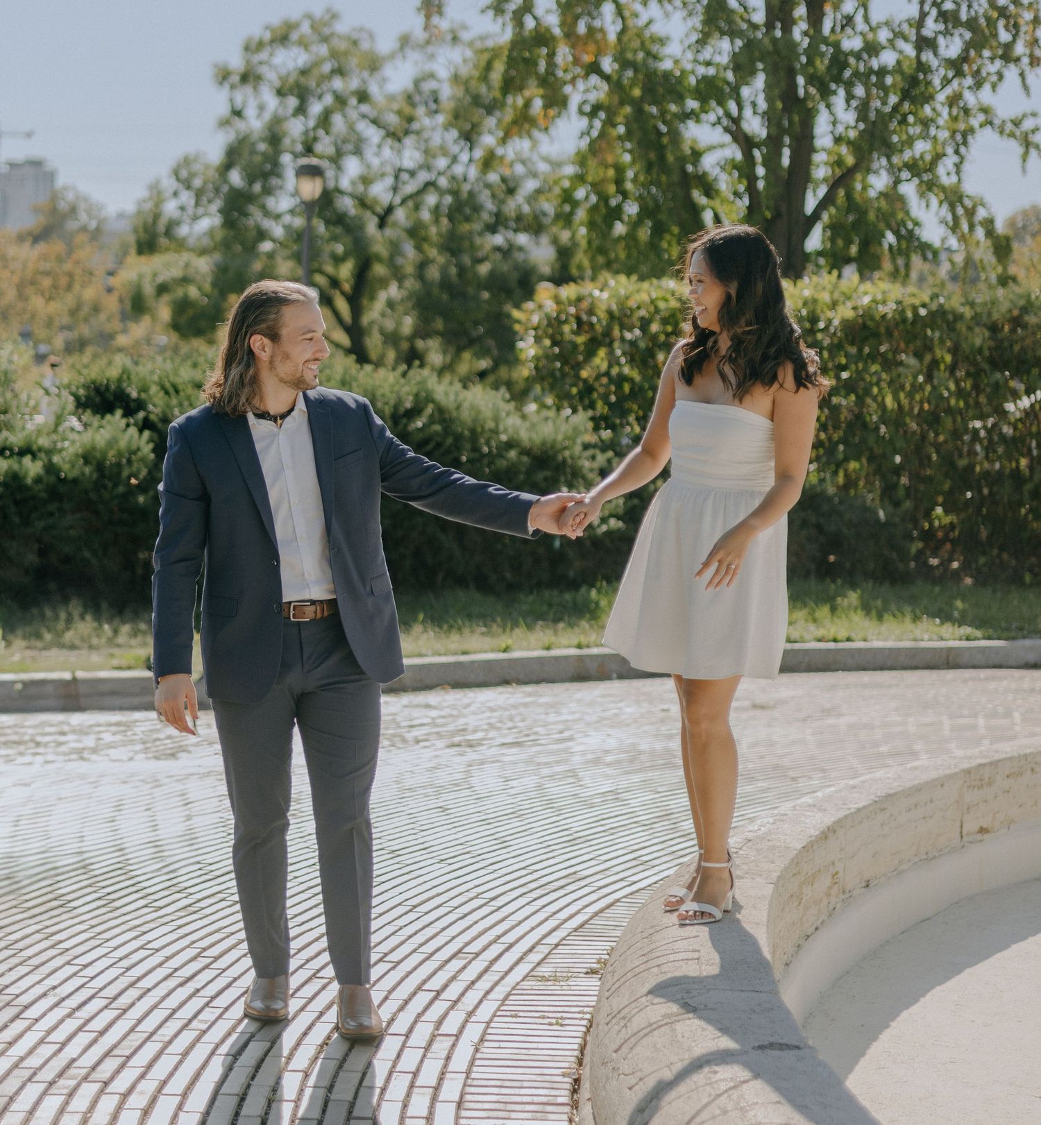 A couple in wedding attire dances and twirls together on white stone steps in a sunny outdoor garden setting.