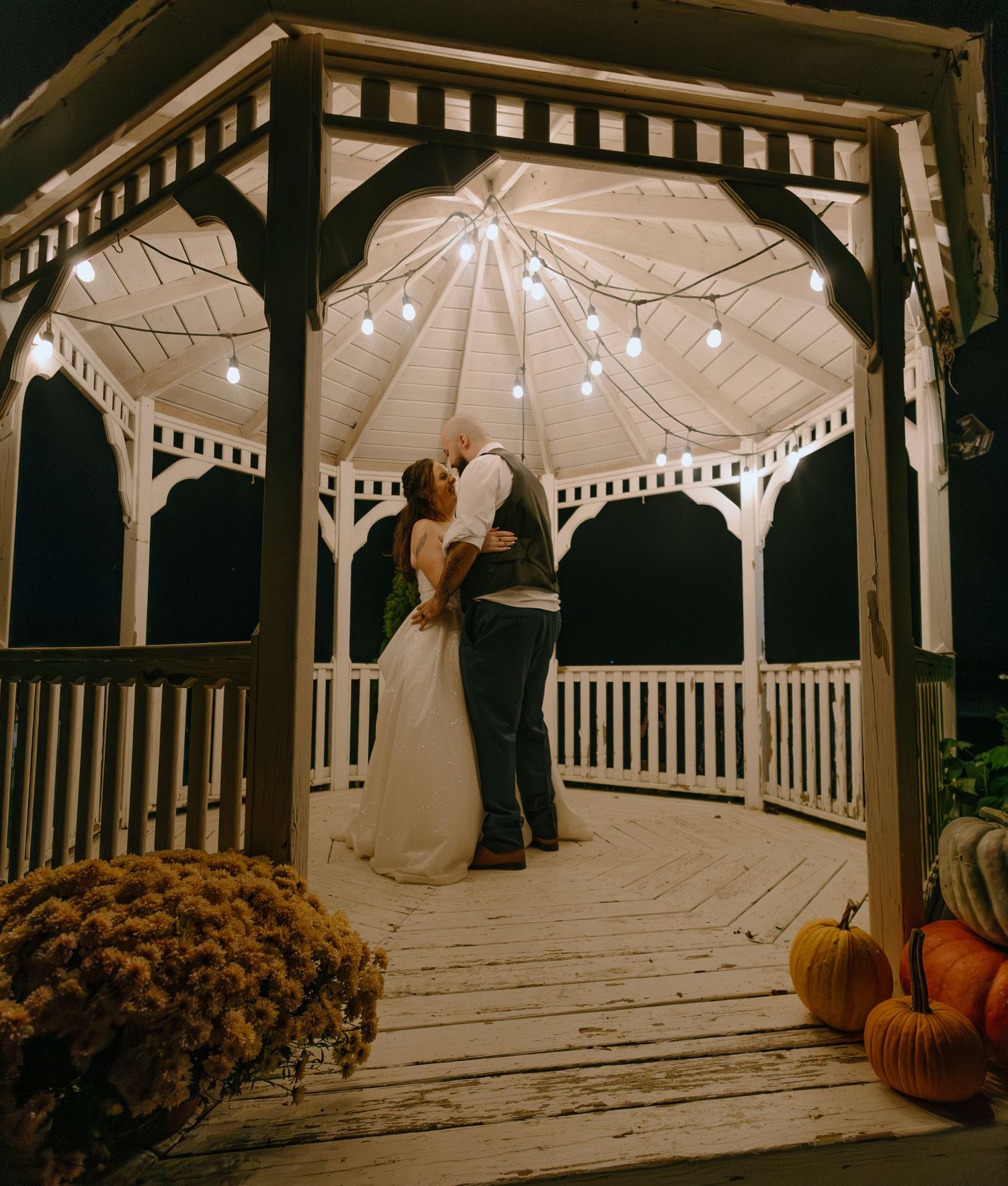 A white wooden gazebo decorated with string lights and fall pumpkins creates a romantic nighttime atmosphere.