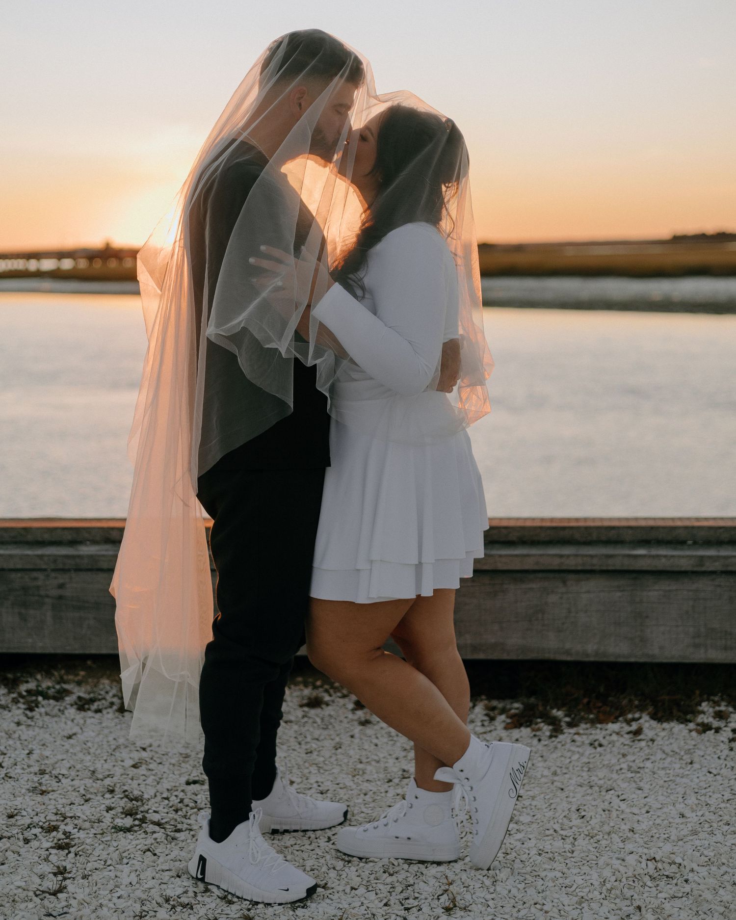 Two figures in white and black outfits standing together by the water at sunset.