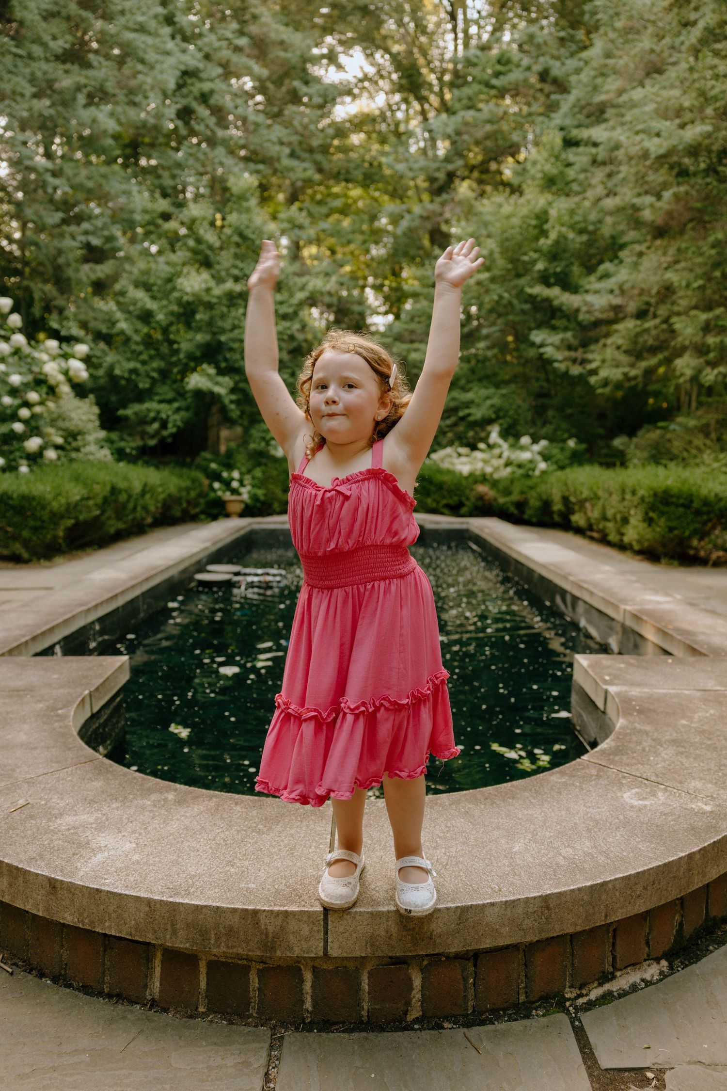 Young child in pink dress plays joyfully on curved concrete wall near water feature.