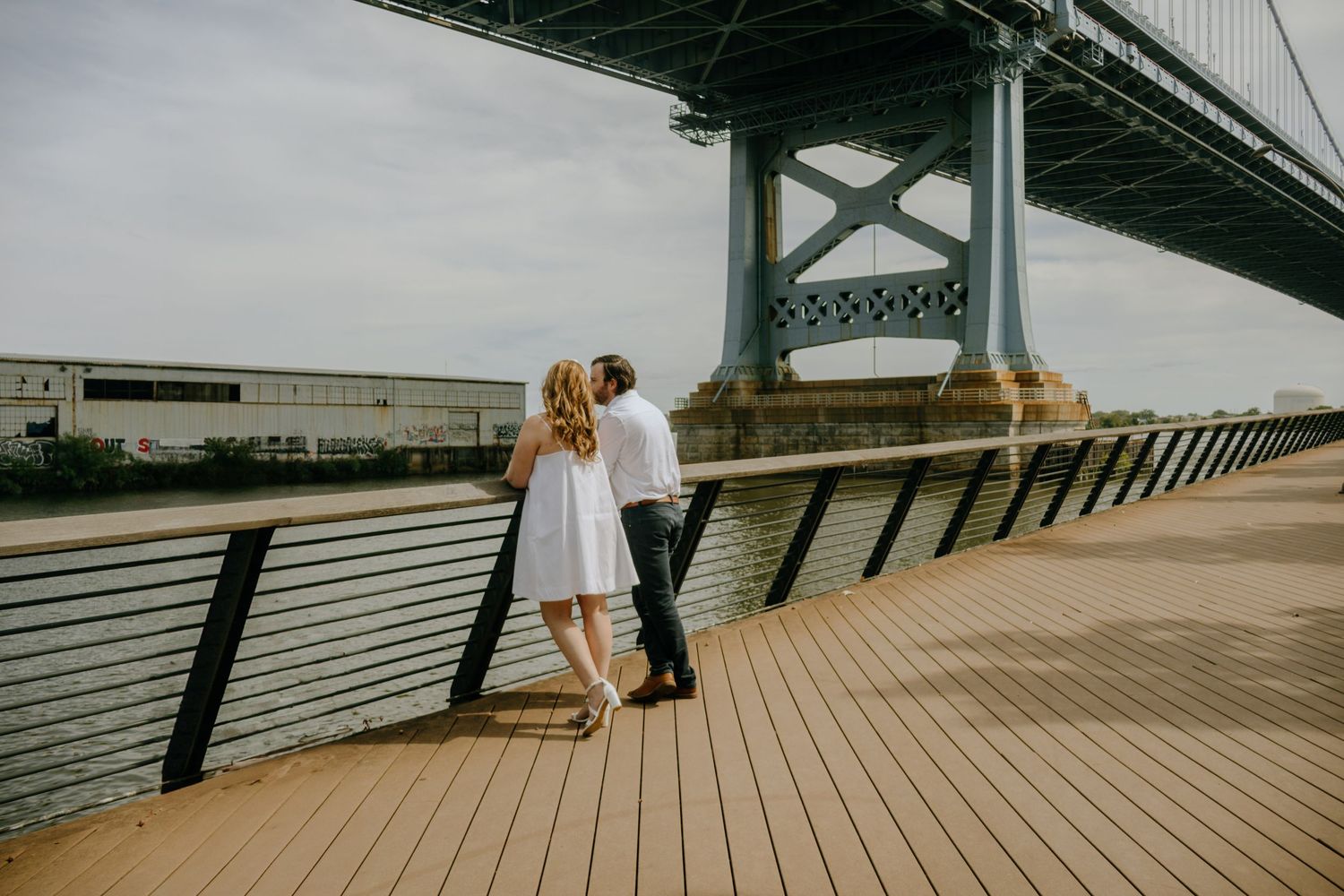 A couple shares a romantic moment on a wooden pier beneath a towering suspension bridge on an overcast day.