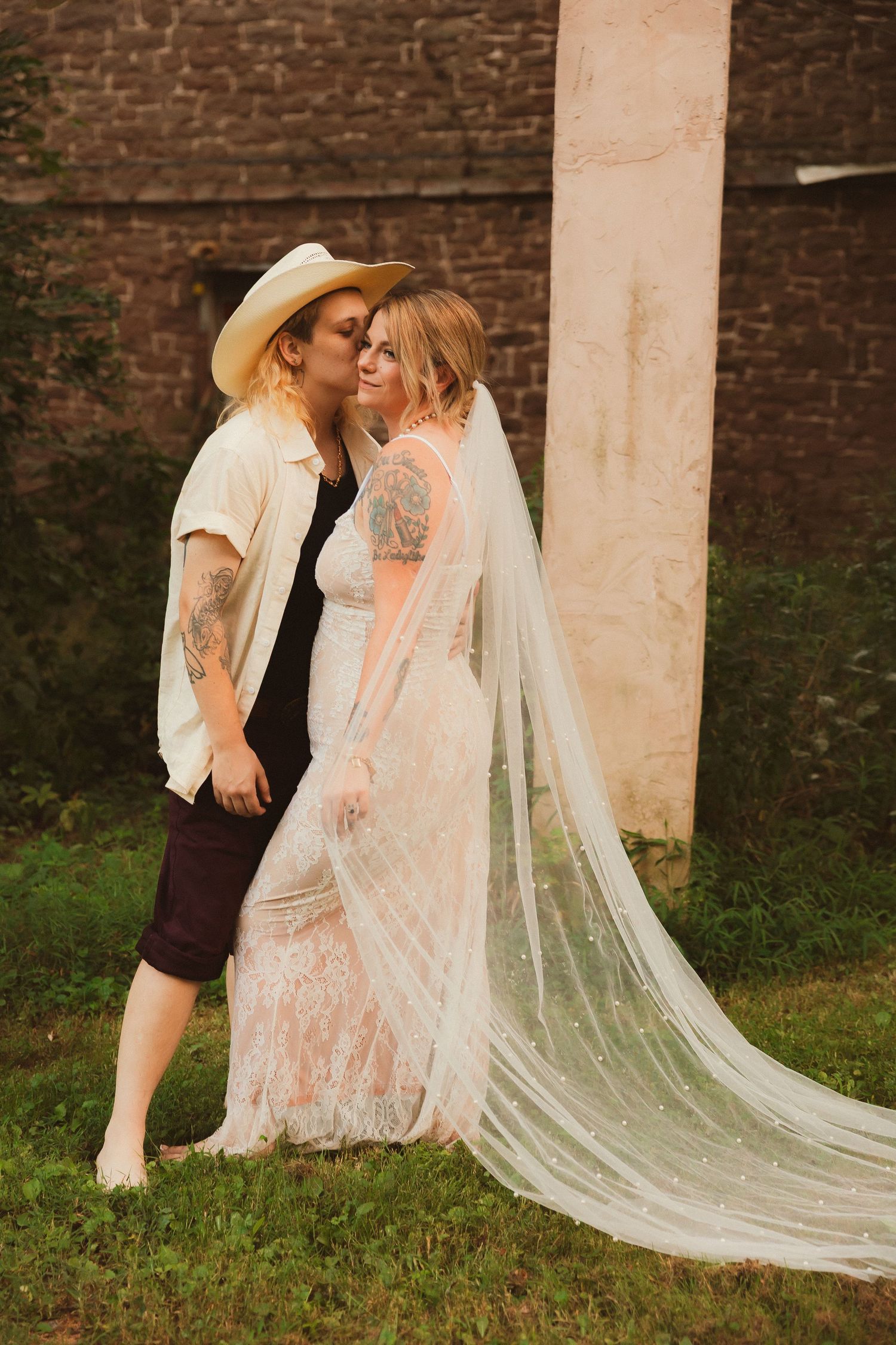A romantic couple posing against a brick wall with a flowing wedding veil in warm sunlight.