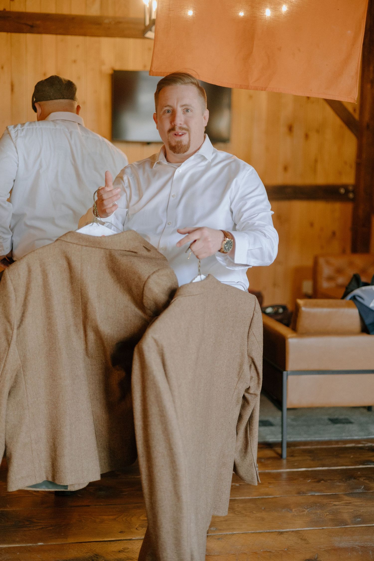 A person in a white dress shirt adjusts formal attire while getting ready in a rustic wooden interior.