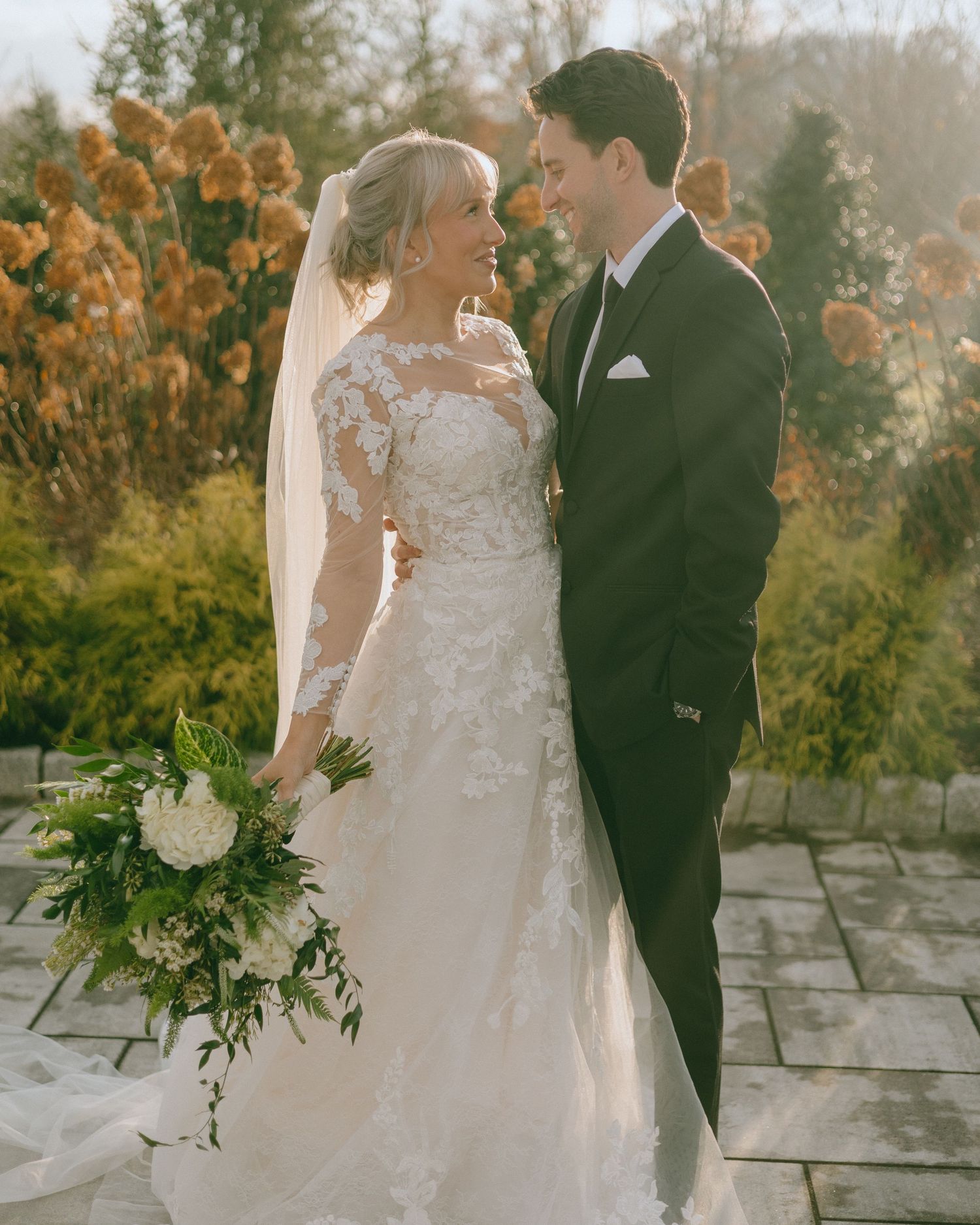 A romantic wedding couple shares an intimate moment at sunset among fall foliage with the bride holding a white bouquet.