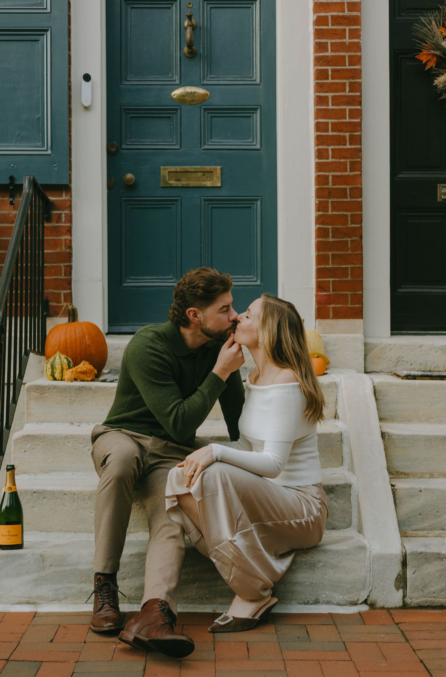 A couple sits together on white stone steps in front of teal-colored doors and brick walls with autumn pumpkin decor.