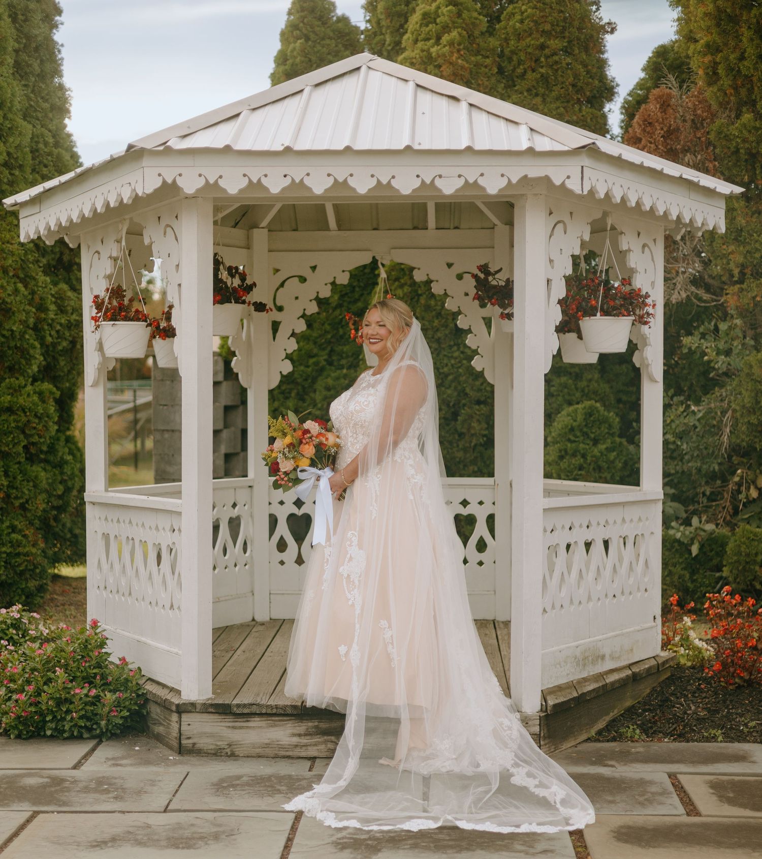 A bride poses in an elegant white gazebo decorated with hanging flowers and ornate trim details.