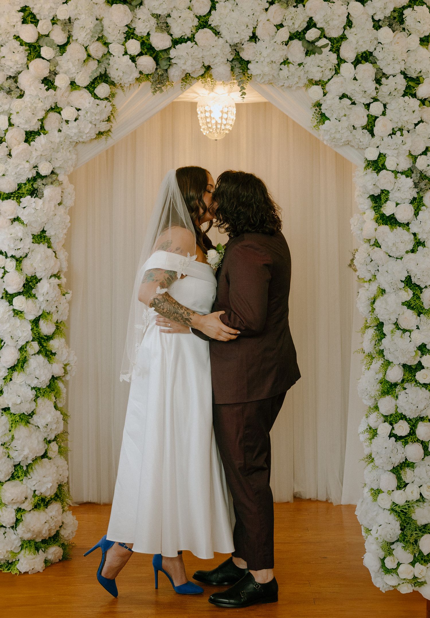 A couple dances under an elegant white floral arch with crystal chandelier at their wedding celebration.