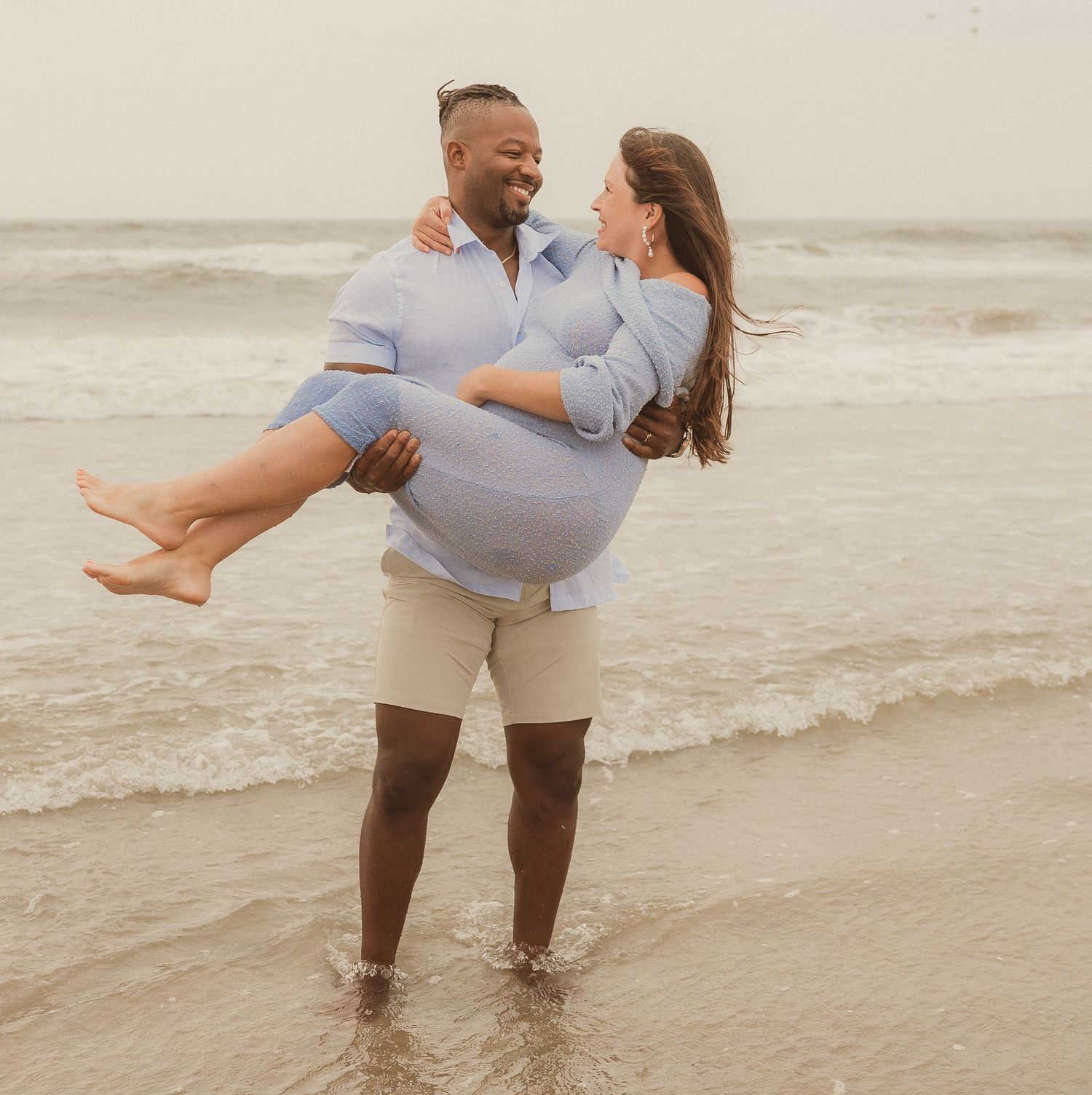 Playful couple embracing and twirling in the shallow ocean surf while wearing casual clothing.