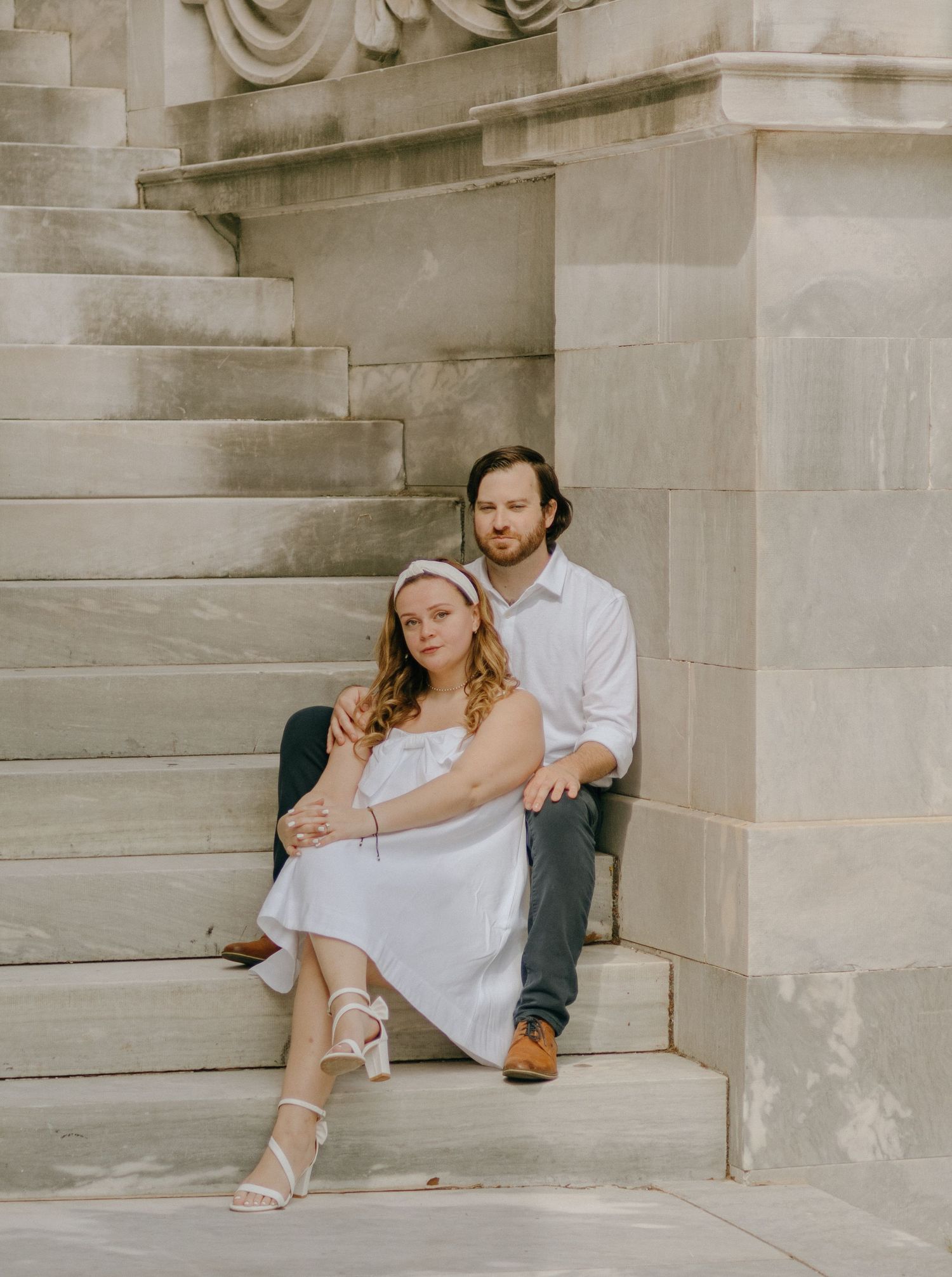 A romantic couple sits together on stone steps in a tender embrace against an architectural backdrop.