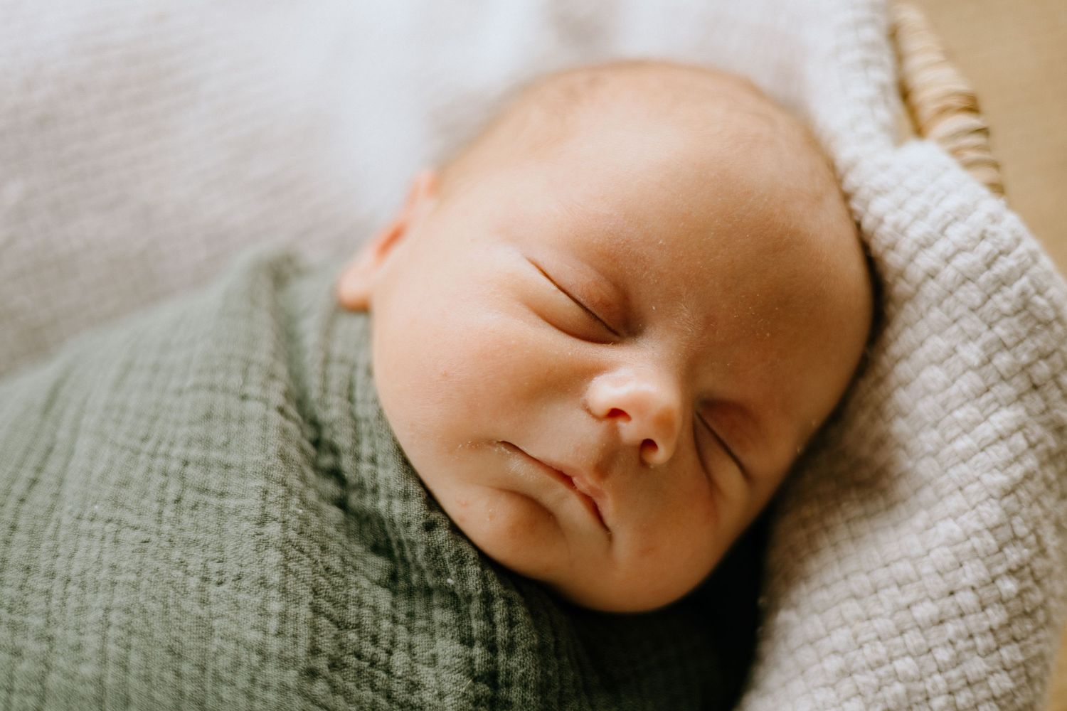 A sleeping newborn baby rests peacefully on textured white bedding in soft natural light.