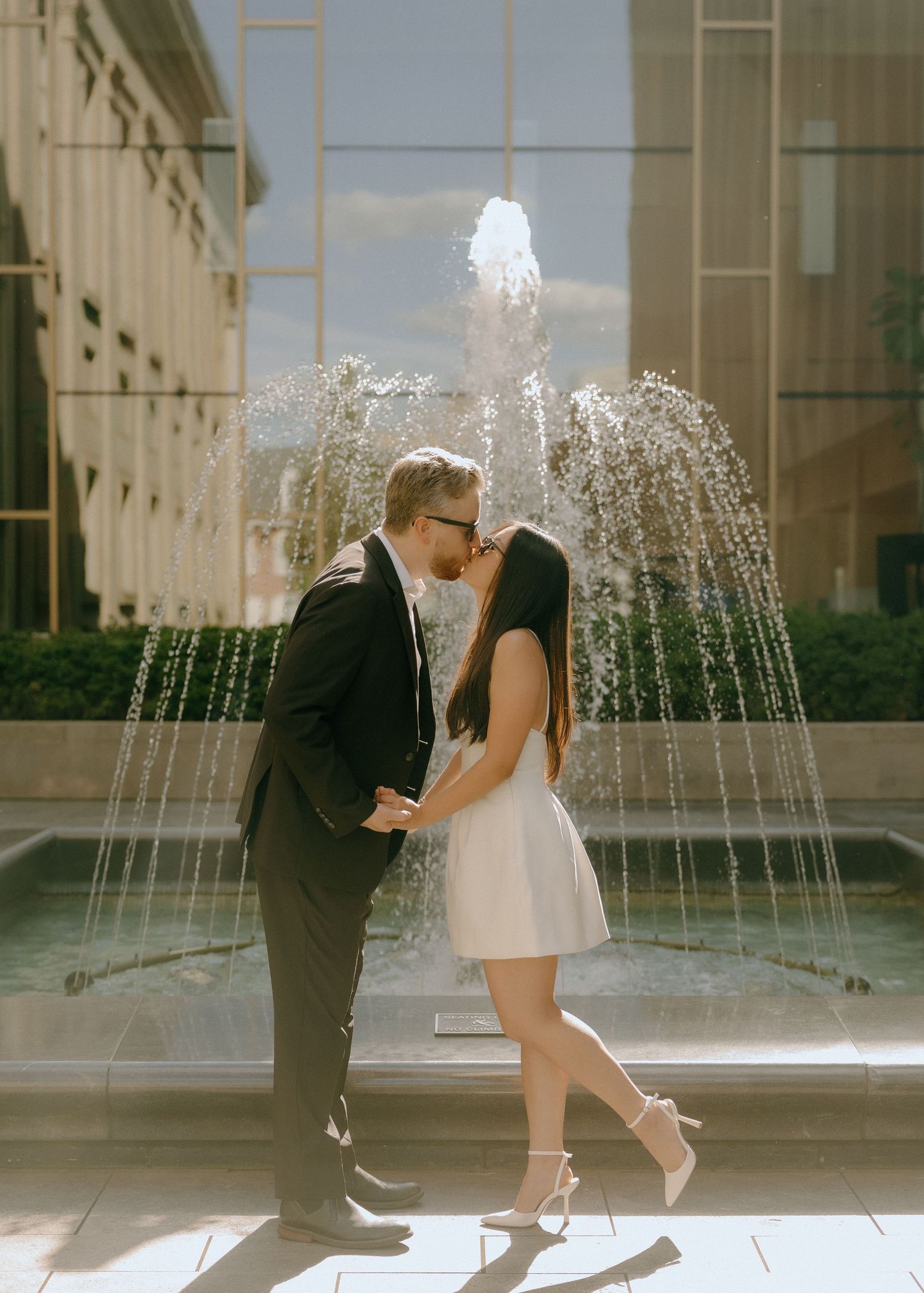 A romantic couple shares intimate moments by a fountain in an elegant outdoor setting.